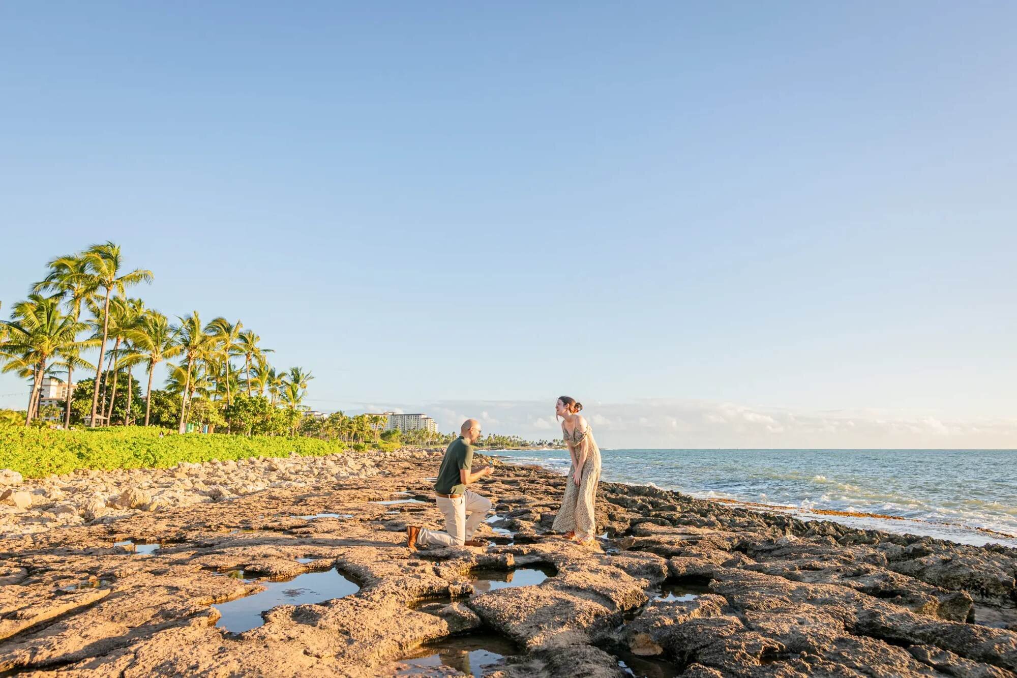 Proposal Photographer Oahu — Surprise Proposal at Ko Olina Beach A man proposing on one knee on lava rocks at Ko Olina beach on Oahu with palm trees and turquoise ocean in the background