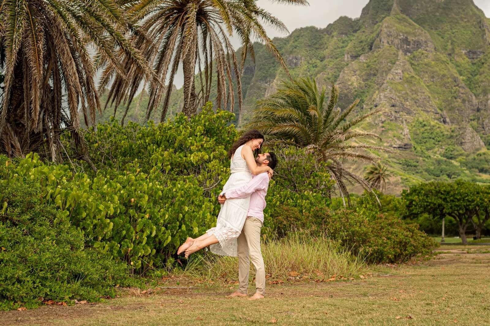 A couple on a grassy lawn, the man lifting the woman in a white lace dress, with palm trees and jagged green mountains behind them