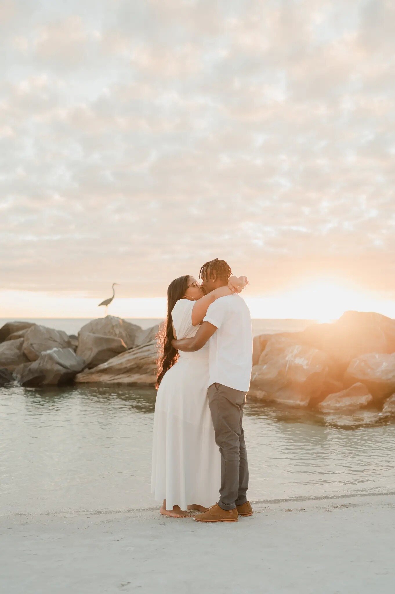 A couple sharing a kiss on a white sand beach at golden sunset in Orlando, Florida, with large rocks, calm water, and a heron perched in the background