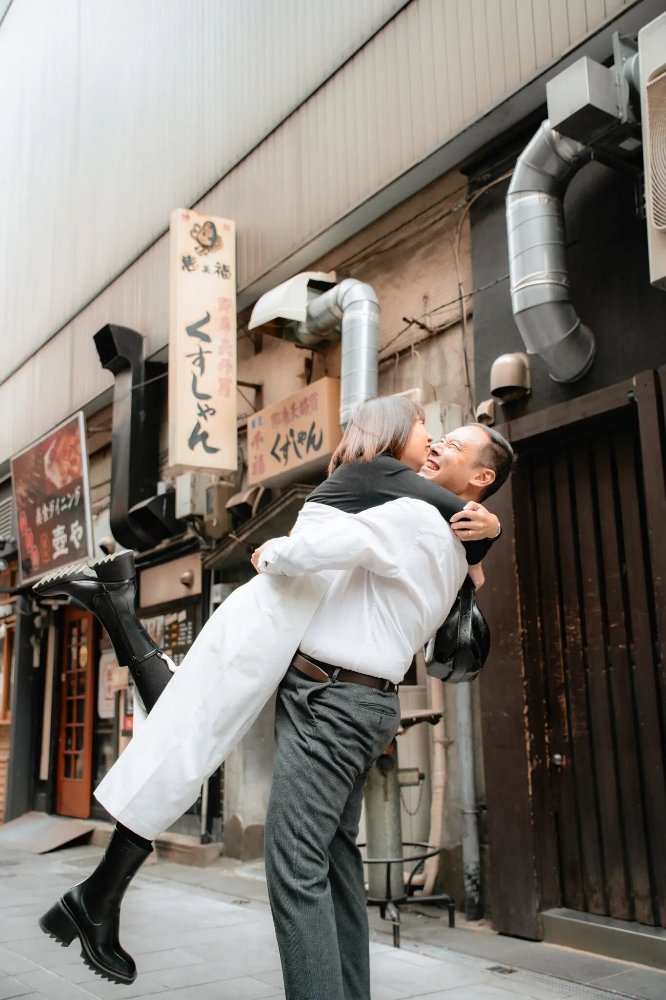 A laughing man lifts a woman in black boots off the ground in a narrow Fukuoka street lined with Japanese restaurant signs and metal ducts