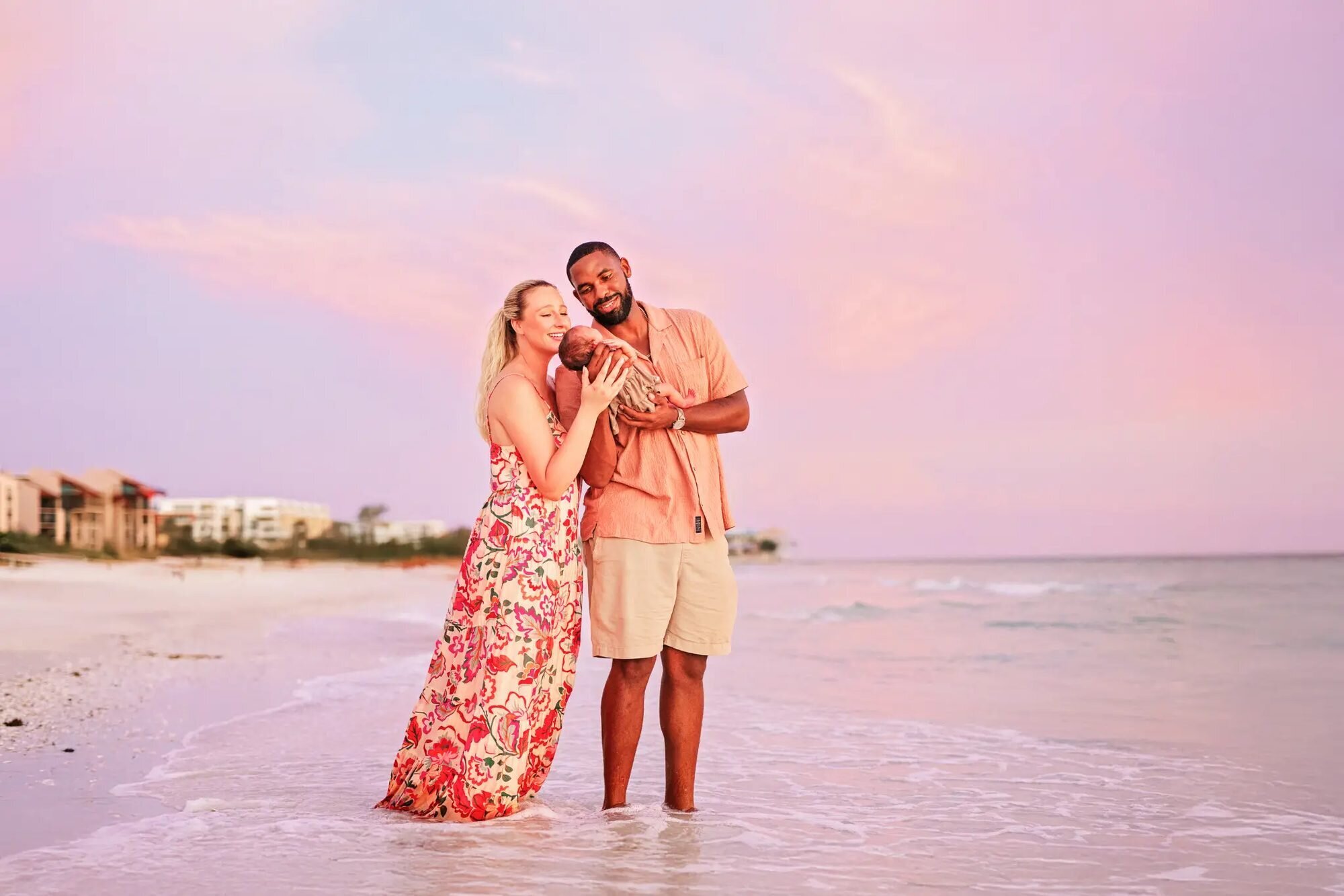 New parents cradling their newborn baby while standing in the shallow Gulf waters at Clearwater Beach, Tampa, during a pink and purple sunset — captured by a Dreamgraphy Tampa family photographer