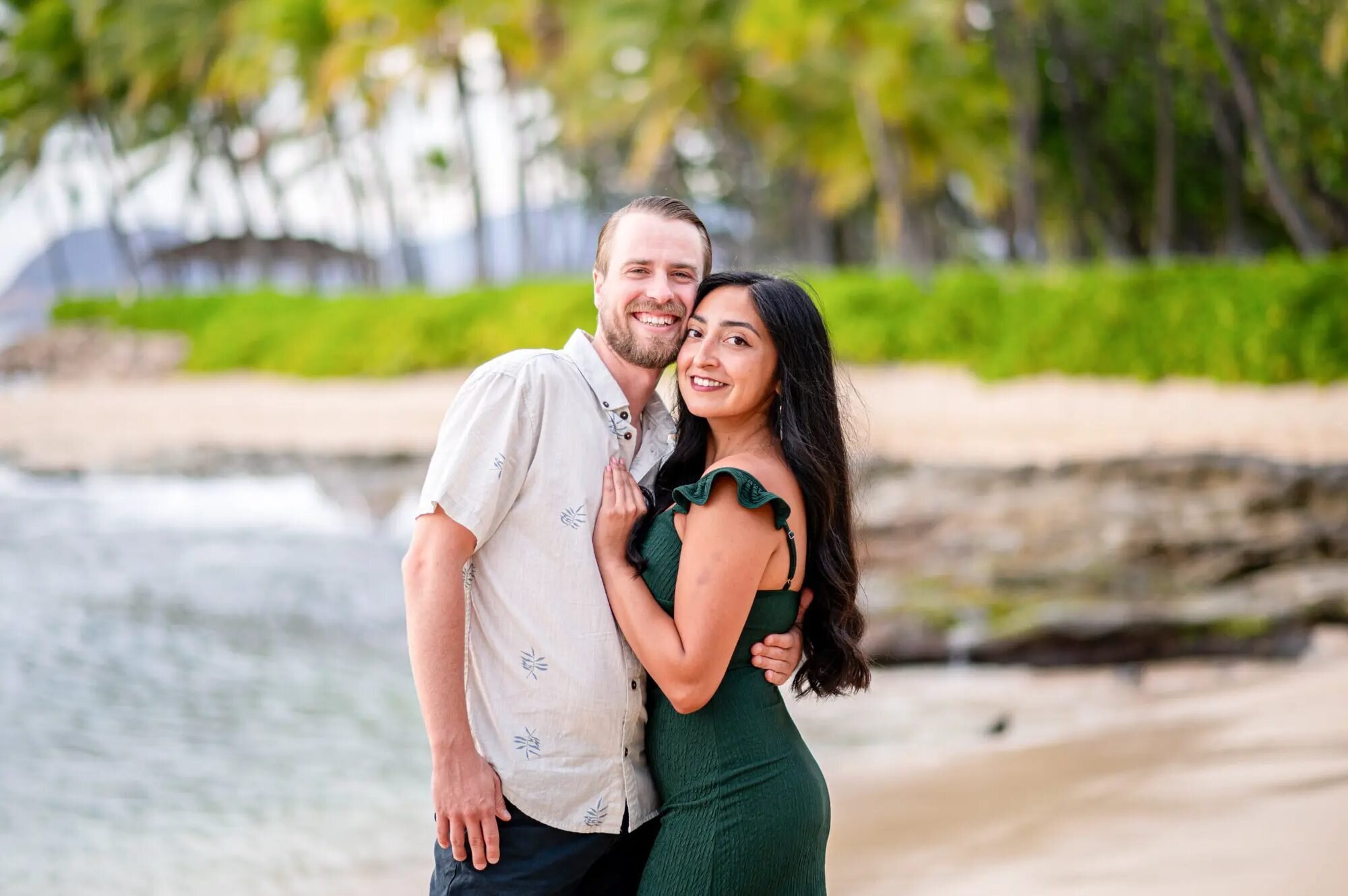 A smiling couple embracing on the sandy shore at Ko Olina Beach on Oahu with lush green tropical foliage and calm water in the background