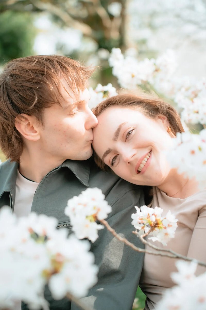 A man kisses a smiling woman on the forehead, framed by white blossoms in soft focus, at Maizuru Park in Fukuoka.