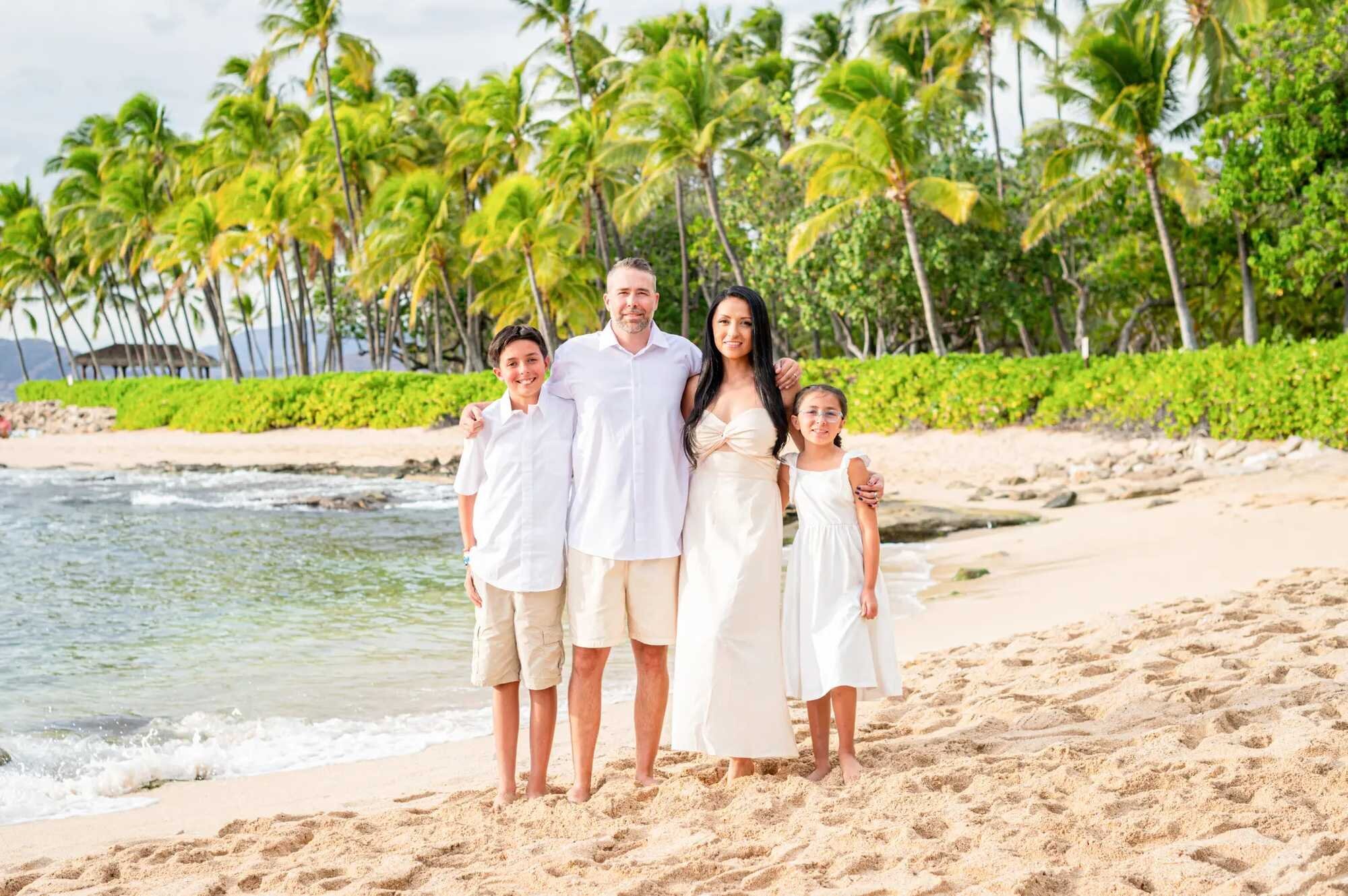 Oahu Photographer — Family Session at Ko Olina Beach A family of four in white and cream outfits posing together on the sandy shore at Ko Olina Beach on Oahu with lush green palm trees and calm turquoise water behind them