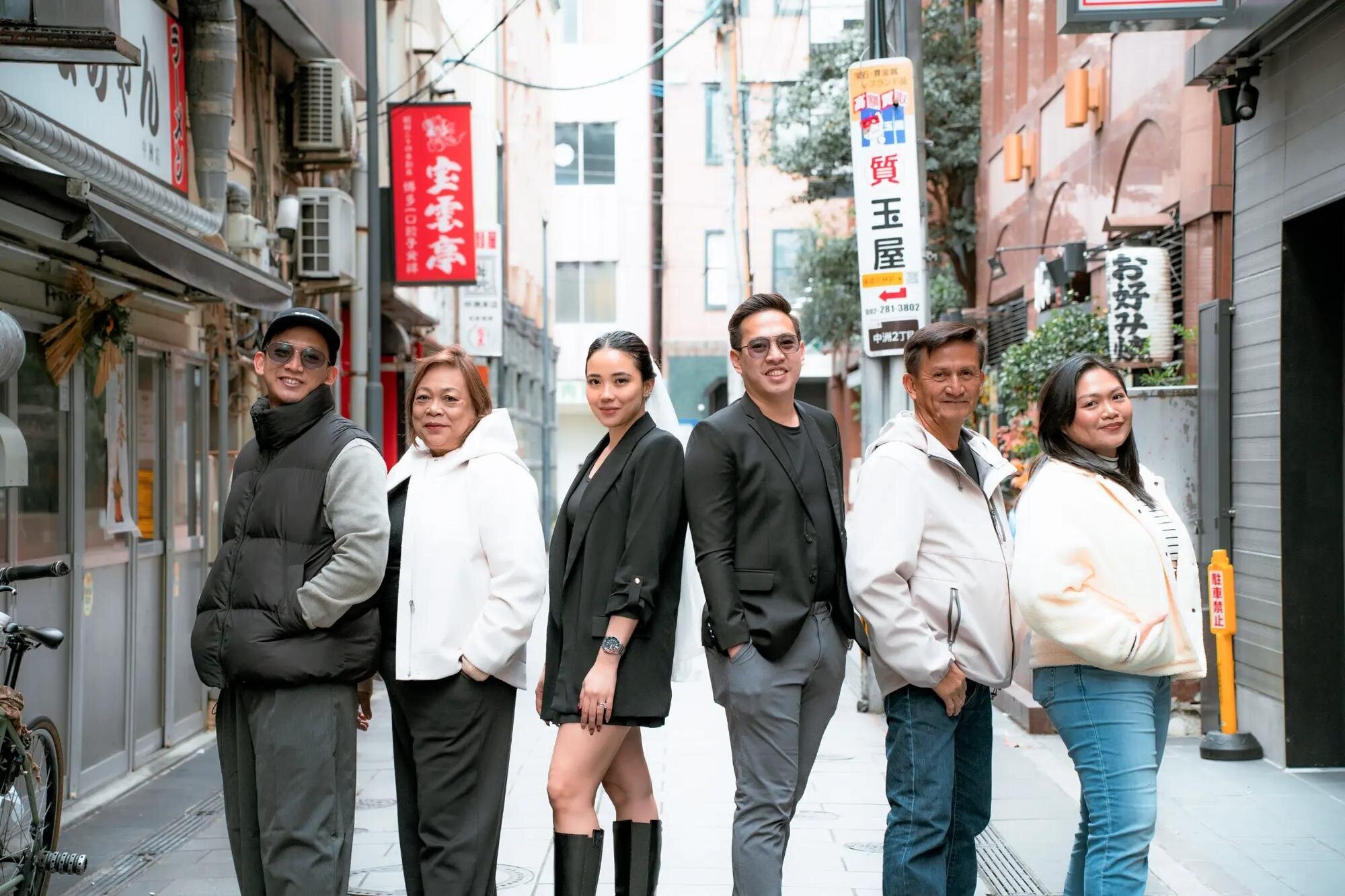  A stylish multigenerational family posing together on a narrow Nakasu alley lined with Japanese shop signs in Fukuoka. A relaxed, editorial-style family photo session captured by Dreamgraphy.