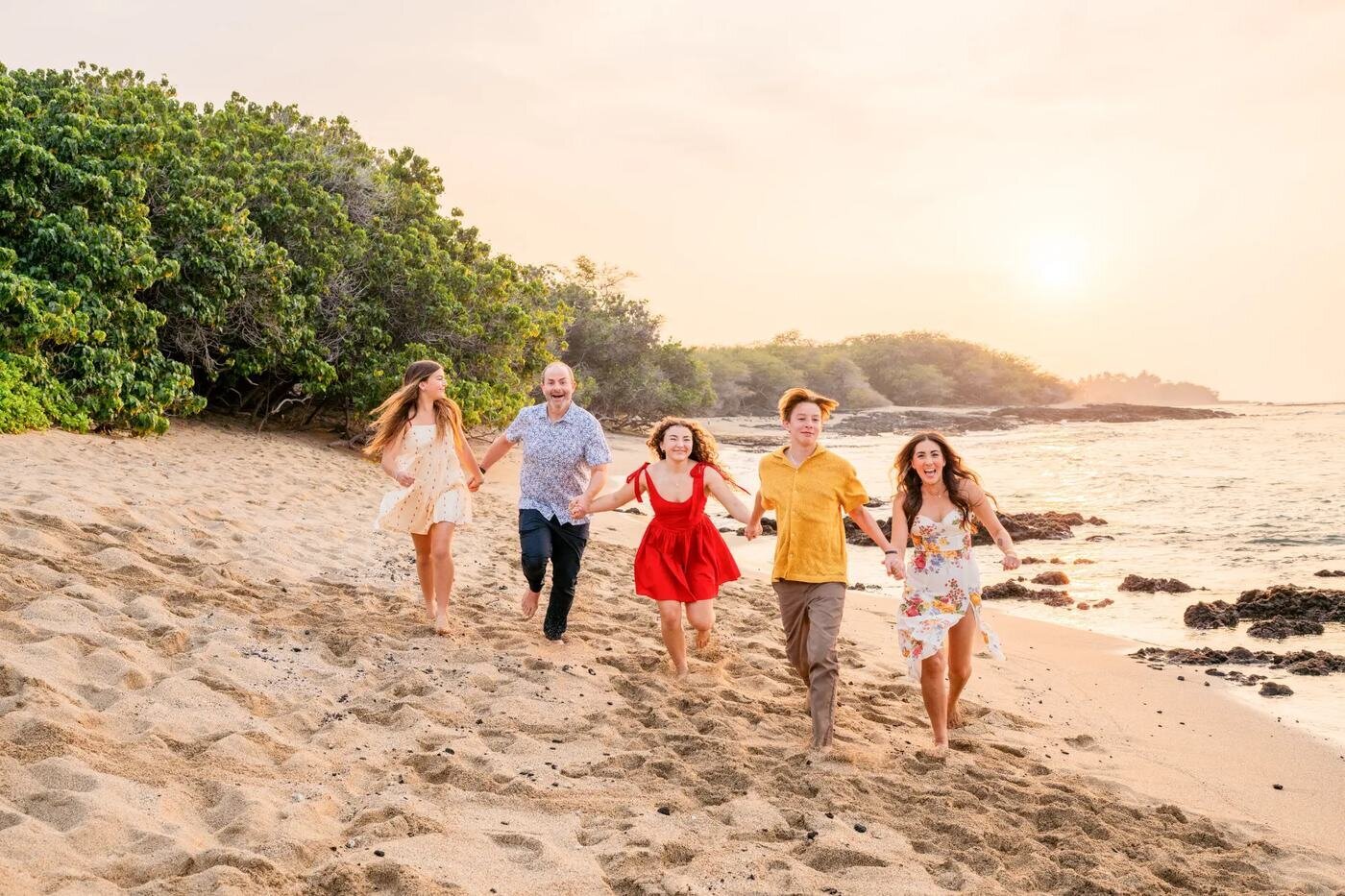 A family of five laughing and running together along a Big Island beach at sunset with lush trees and ocean in the background
