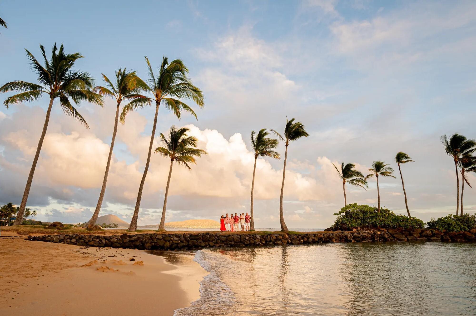 Oahu Photographer — Family Session at Waialae Beach at Sunset A family group posing on a stone jetty at Waialae Beach on Oahu surrounded by tall swaying palm trees with golden sunset light and calm ocean water reflecting the sky