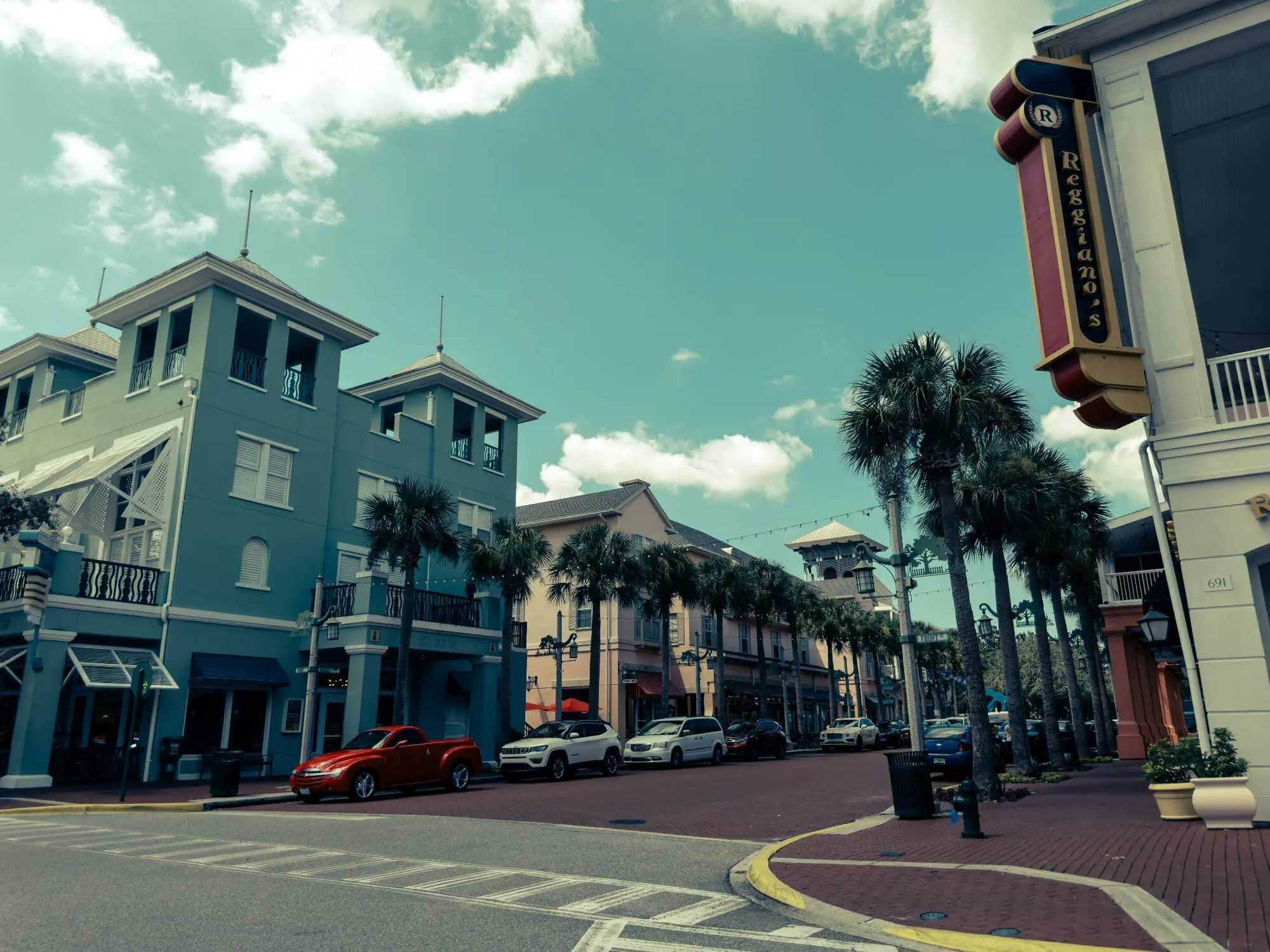 A palm-lined brick street with a two-towered teal building, pastel shopfronts and parked cars in Downtown Celebration, Florida