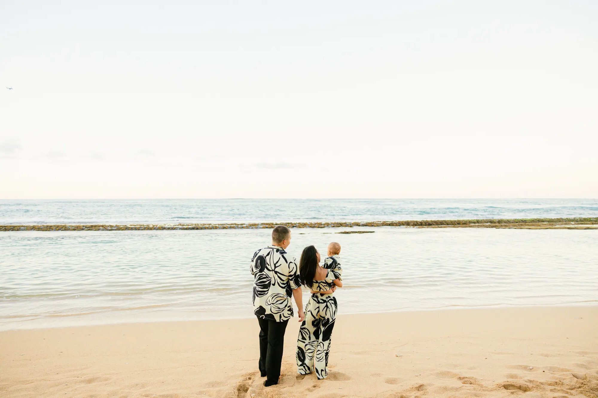 A couple in matching Hawaiian print outfits holding their newborn baby while standing on a sandy Maui beach looking out at the calm ocean