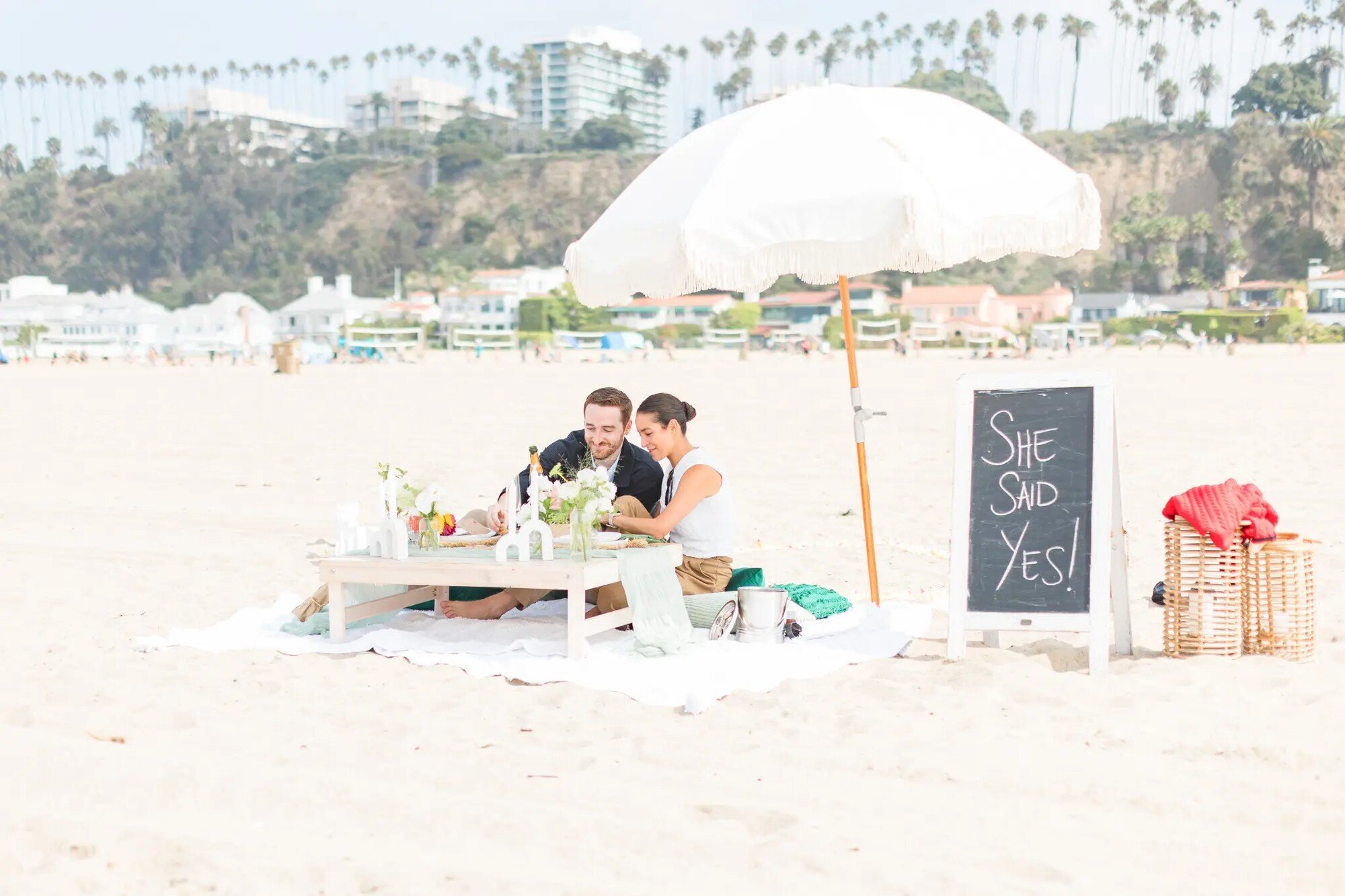 A couple celebrating a surprise beach picnic proposal with a She Said Yes sign — engagement proposal photographer by Dreamgraphy