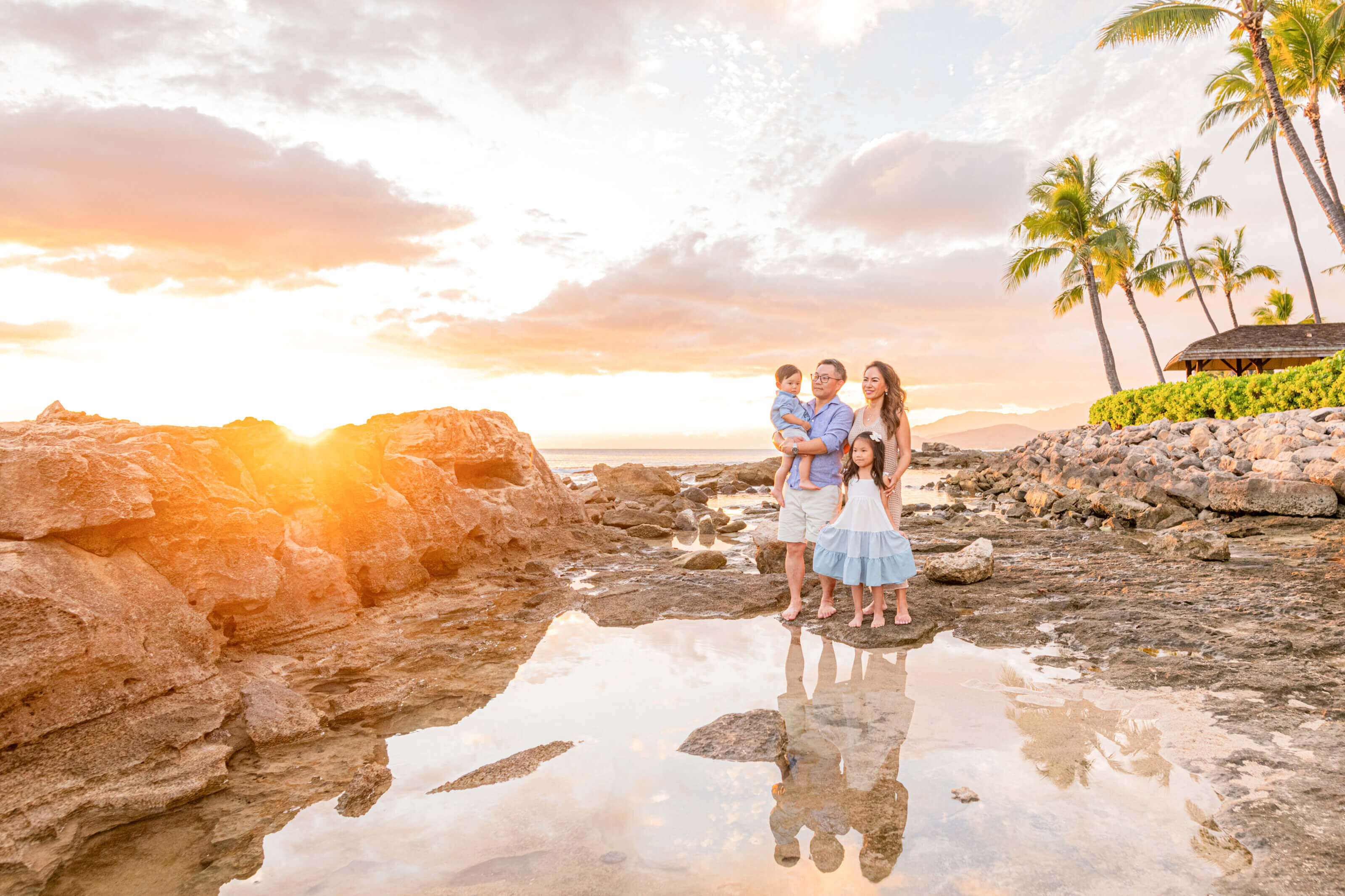 A family of four standing on lava rocks at golden sunset in Hawaii with their reflection in a tidal pool and palm trees glowing in the warm light during a vacation photoshoot