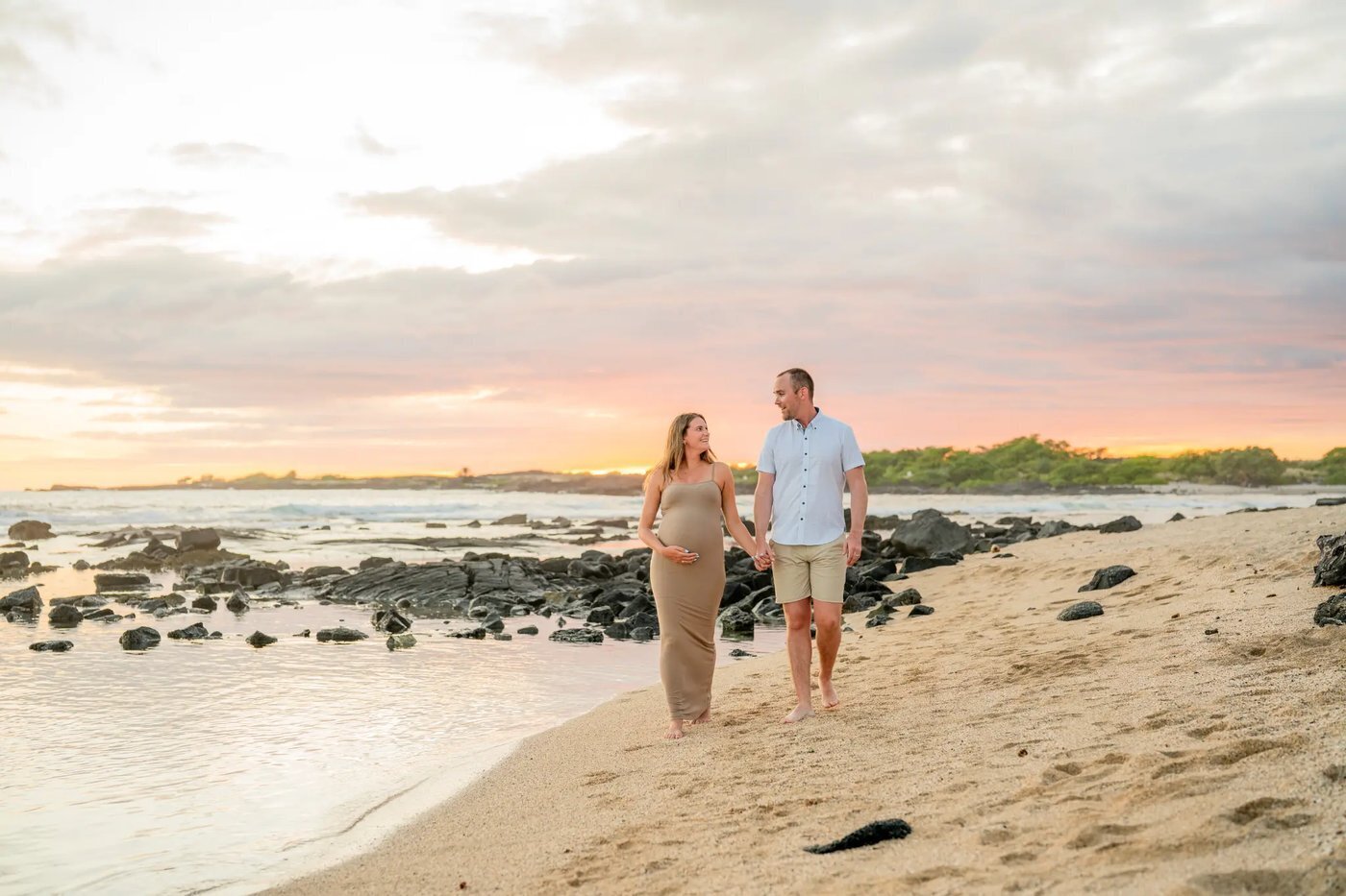 A pregnant woman in a tan dress holding her belly while walking hand in hand with her partner on a Big Island beach at sunset with lava rocks and ocean in the background