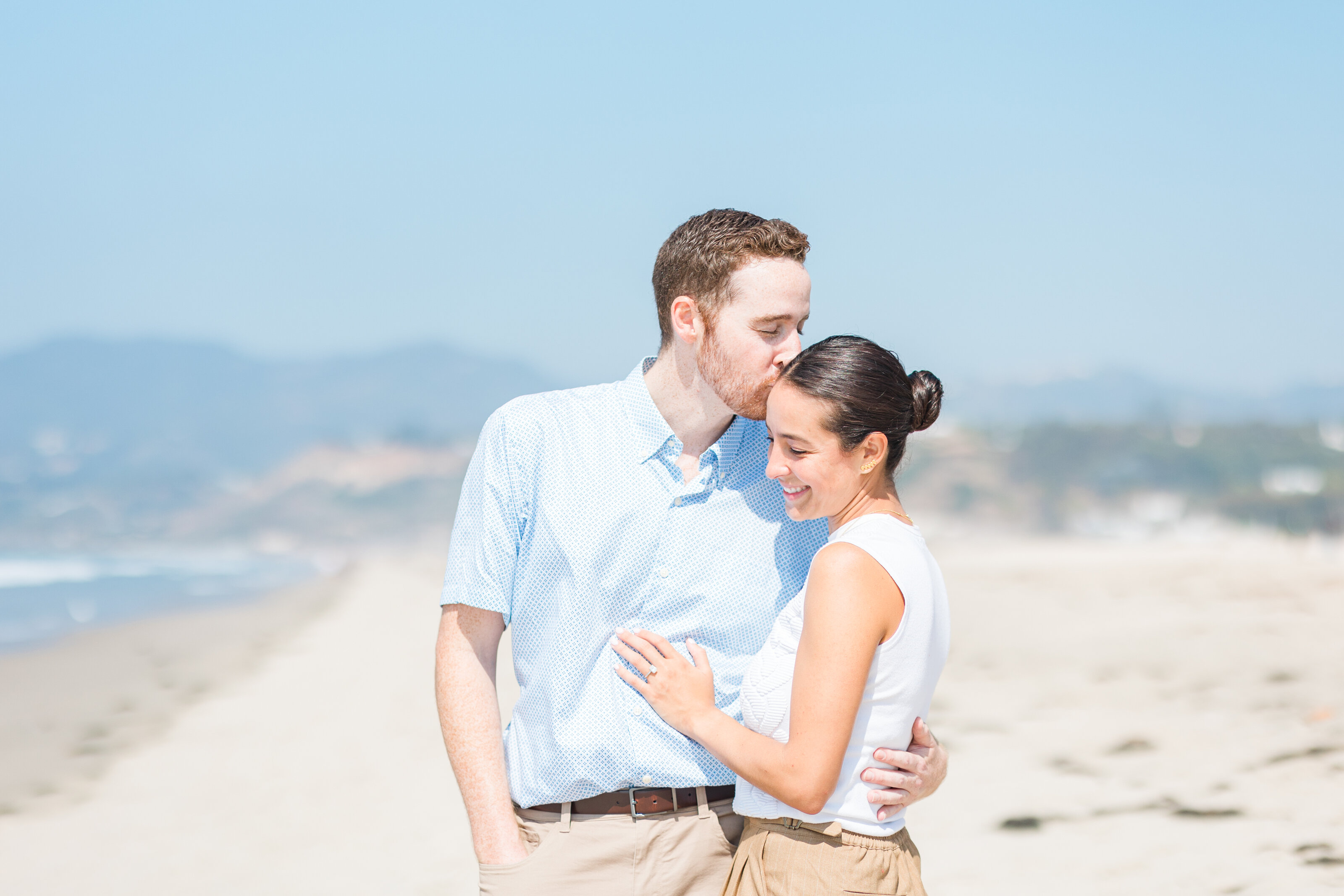 A man kissing the forehead of a smiling woman as they embrace on a sunny beach in Los Angeles, with hazy hills and the ocean behind