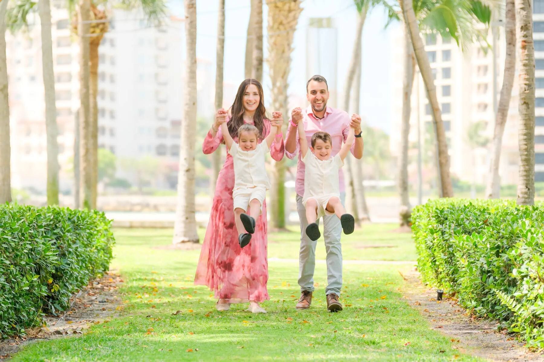miami-photographer-south-pointe-park-session A family of four walks along a grass path between hedges at South Pointe Park, the parents in pink swinging two small boys by their arms