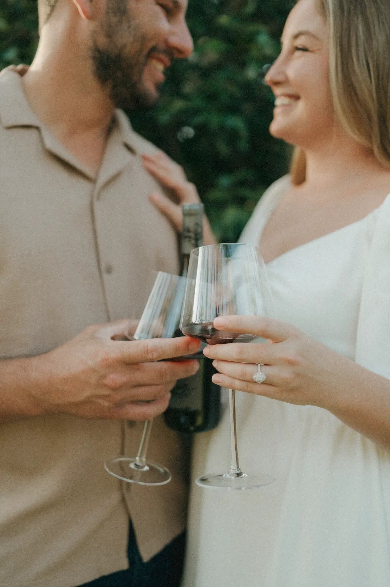 An engaged couple smiling and toasting with wine glasses during a romantic engagement photography session in a lush green garden in Orlando, Florida, with the woman's diamond ring visible
