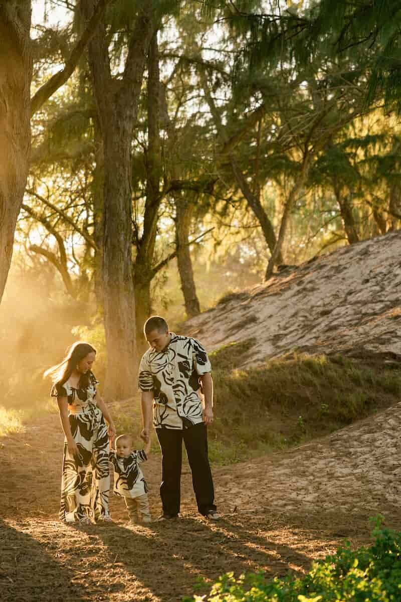 maui-family-photographer-dreamgraphy-golden-hour-ironwood A couple standing on a sandy hillside holding their baby hands and smiling during a family vacation photography session in Hawaii, with tall trees and dramatic green mountain cliffs in the background