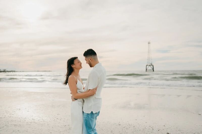A couple embracing and laughing on the white sand beach at Clearwater Beach, Tampa, with a pier and calm Gulf waters in the background — captured by a Dreamgraphy Tampa photographer