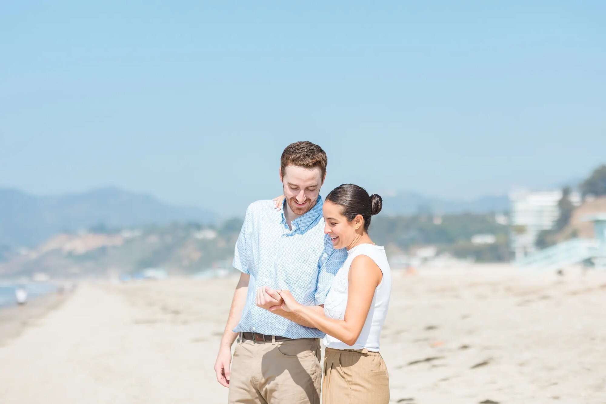 Los Angeles couples photographer capturing a couple walking hand in hand along Santa Monica Beach, smiling and looking at each other with the ocean and cliffs in the background