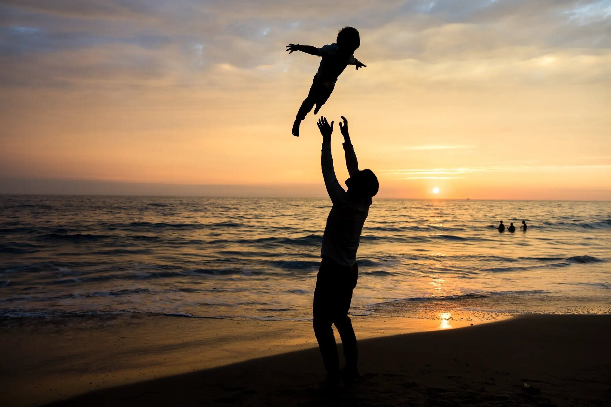Los Angeles family photographer capturing a father tossing his young child high into the air on Santa Monica Beach at sunset, with the ocean and golden sky silhouetted behind them
