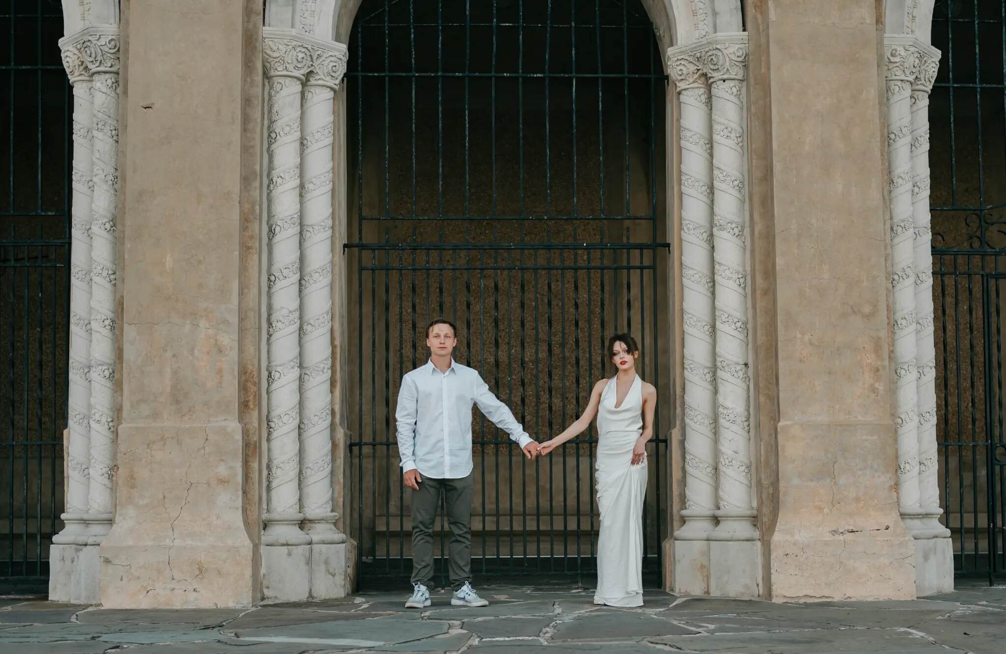 A couple holding hands and posing in front of an ornate stone archway with decorative columns during a couples photography session in Orlando, Florida, the woman wearing an elegant white gown