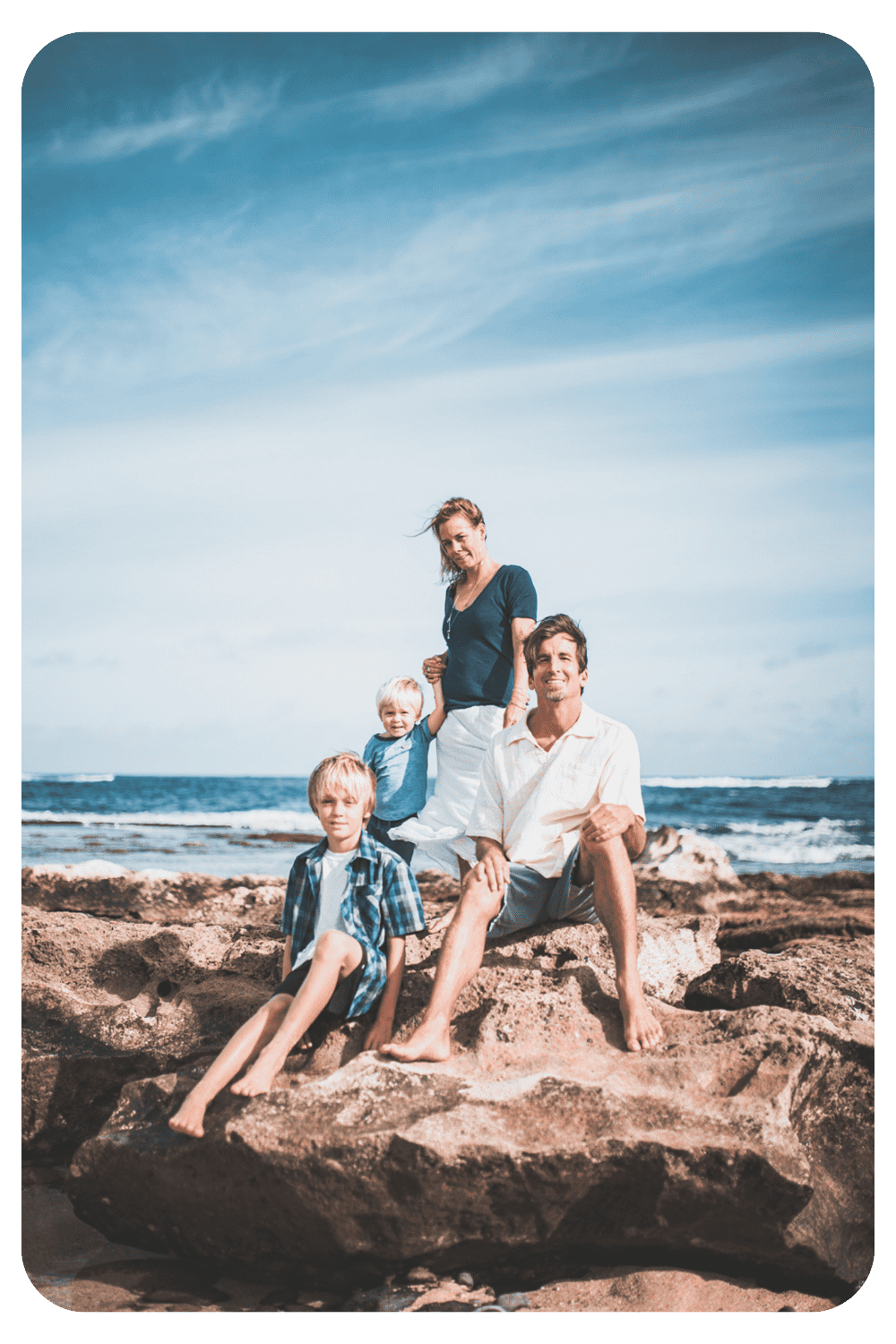 A family of four sitting and standing on volcanic lava rocks along the Maui coastline with the Pacific Ocean and blue sky in the background