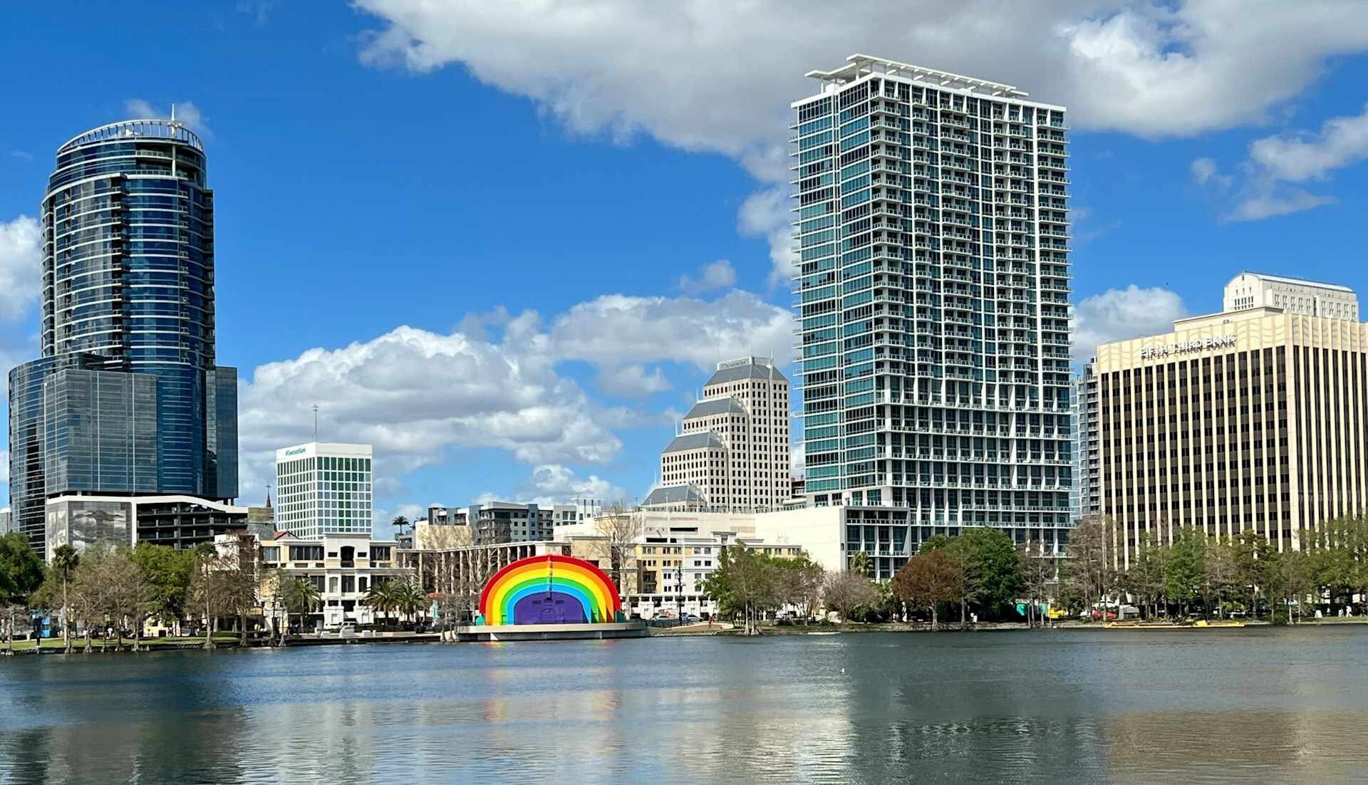 A panoramic view of Lake Eola Park in Orlando, Florida, with the colorful rainbow amphitheater stage reflected in the calm water and the modern glass city skyline rising behind it under a bright blue sky