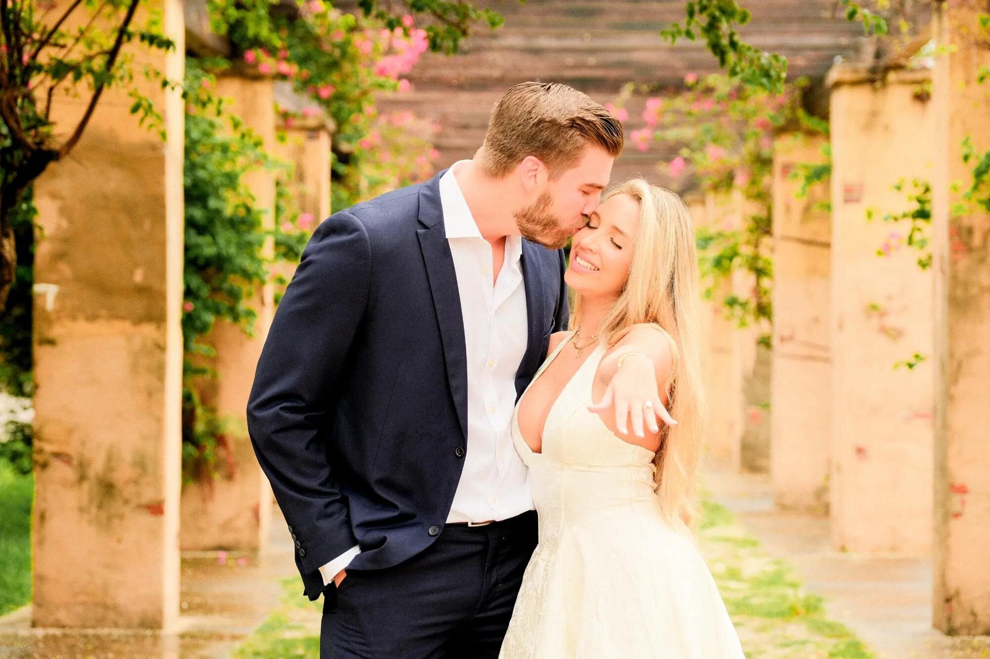 Miami Proposal Photographer | Country Club Prado Engagement Session An engaged couple sharing a tender moment surrounded by flowering vines and stone columns at Country Club Prado, Miami — captured by a Dreamgraphy Miami proposal photographer