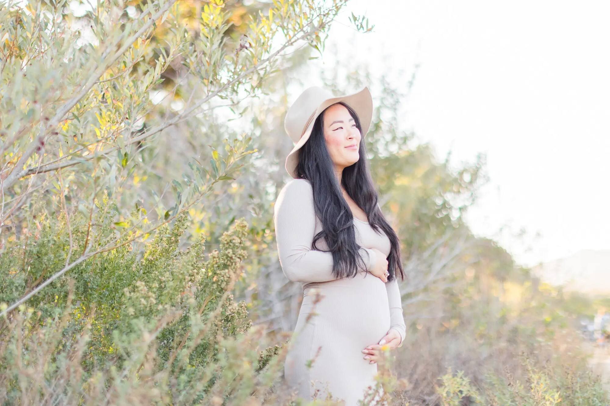 Los Angeles maternity photographer capturing a pregnant woman in a beige dress and wide-brim hat cradling her baby bump while standing among golden wildflowers and olive trees at Malibu Creek State Park