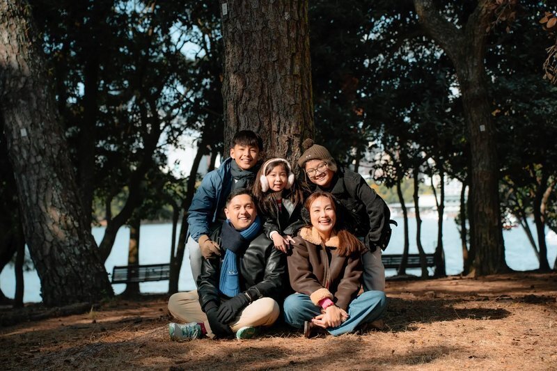 A smiling family of five in winter coats gathers under tall pine trees beside the water at Ohori Park in Fukuoka.