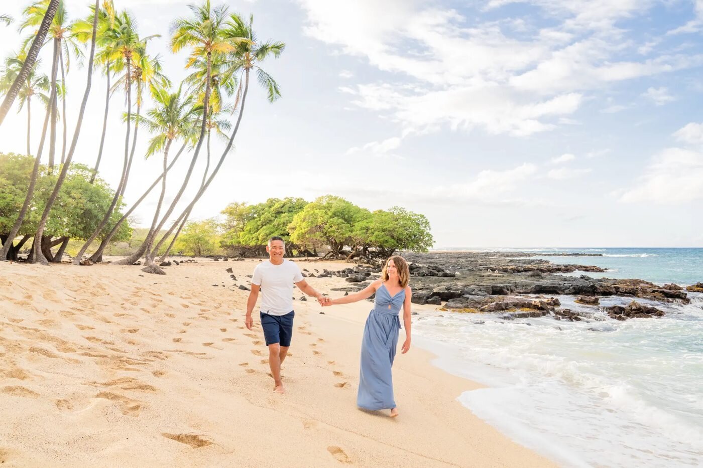 A smiling couple walking hand in hand along a golden sand beach on the Big Island with palm trees and turquoise water behind them