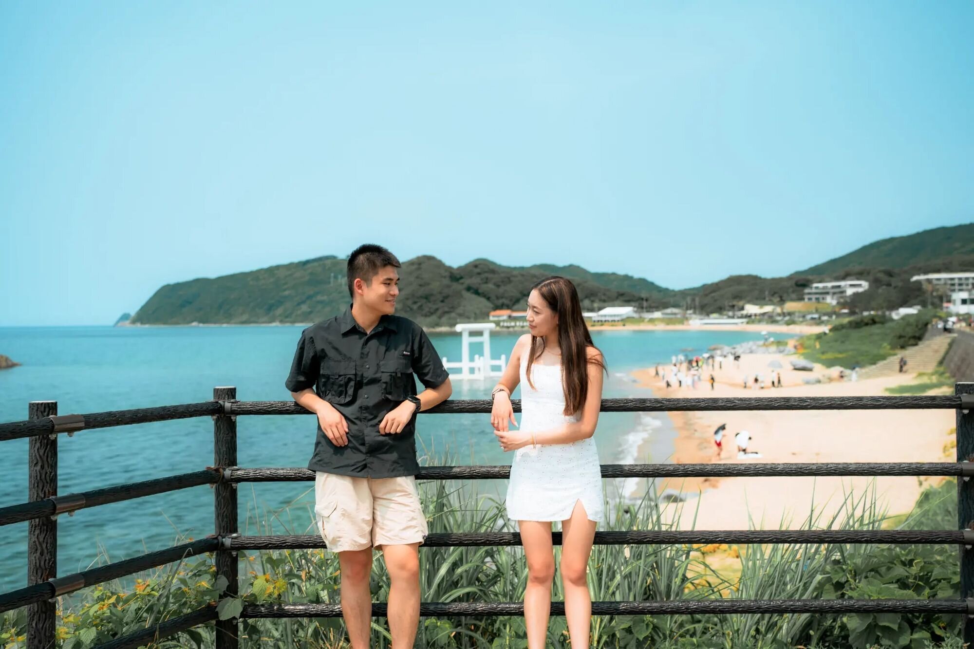 A couple leans on a railing above Futamigaura Beach in Itoshima, with a white torii gate standing in the turquoise sea behind them.