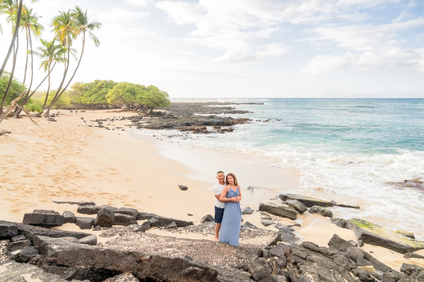 A couple embracing on lava rocks on the Big Island of Hawaii with a wide white sand beach, palm trees, and turquoise ocean stretching behind them
