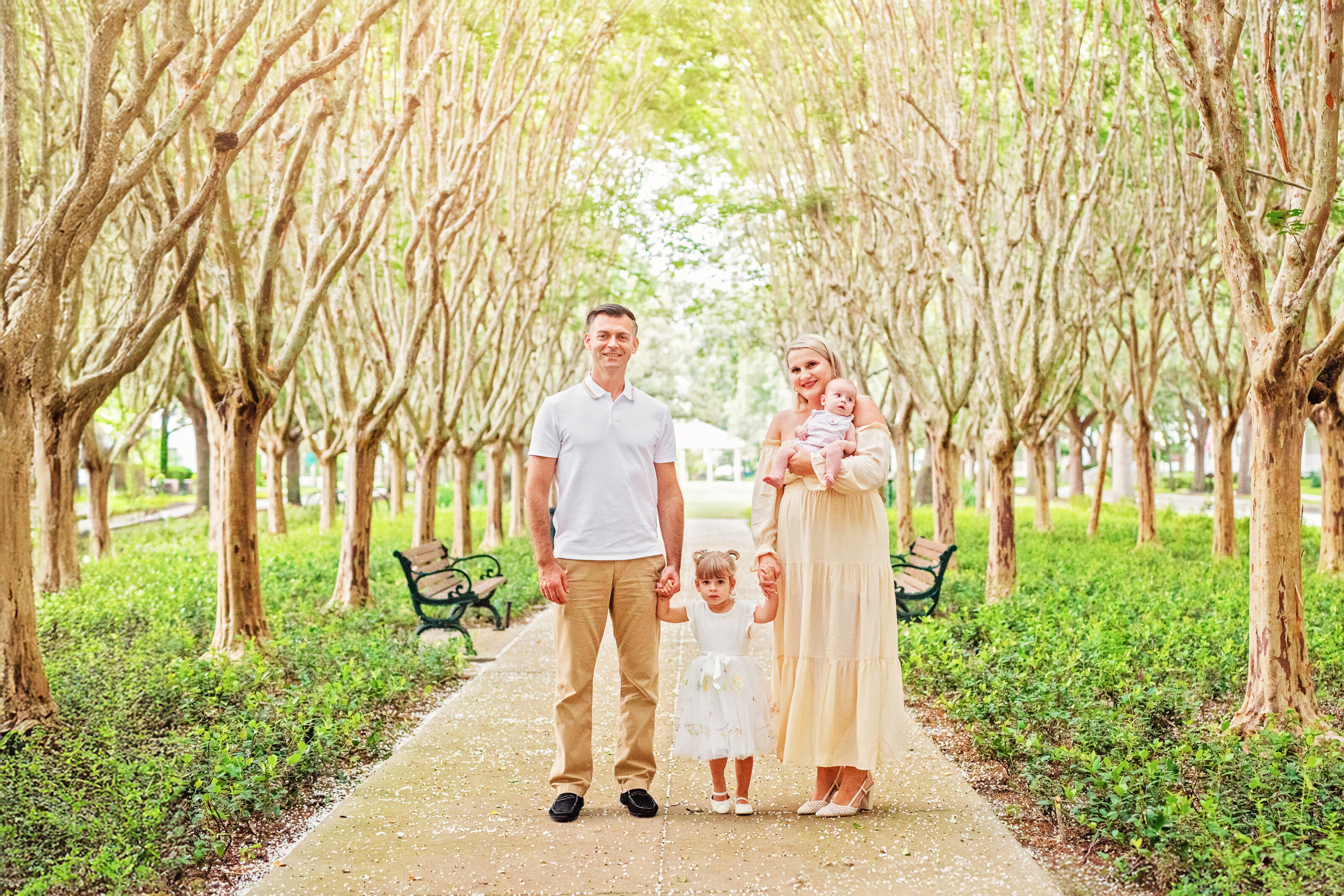 A family of four posing on a tree-lined pathway in Celebration, Florida, with a mother holding a newborn, a toddler daughter between the parents, and lush green gardens and park benches in the background