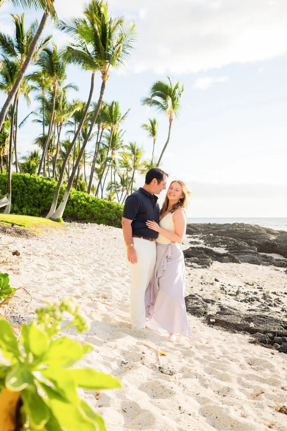 A couple embracing on a white sand beach on the Big Island of Hawaii surrounded by tall palm trees and soft golden light
