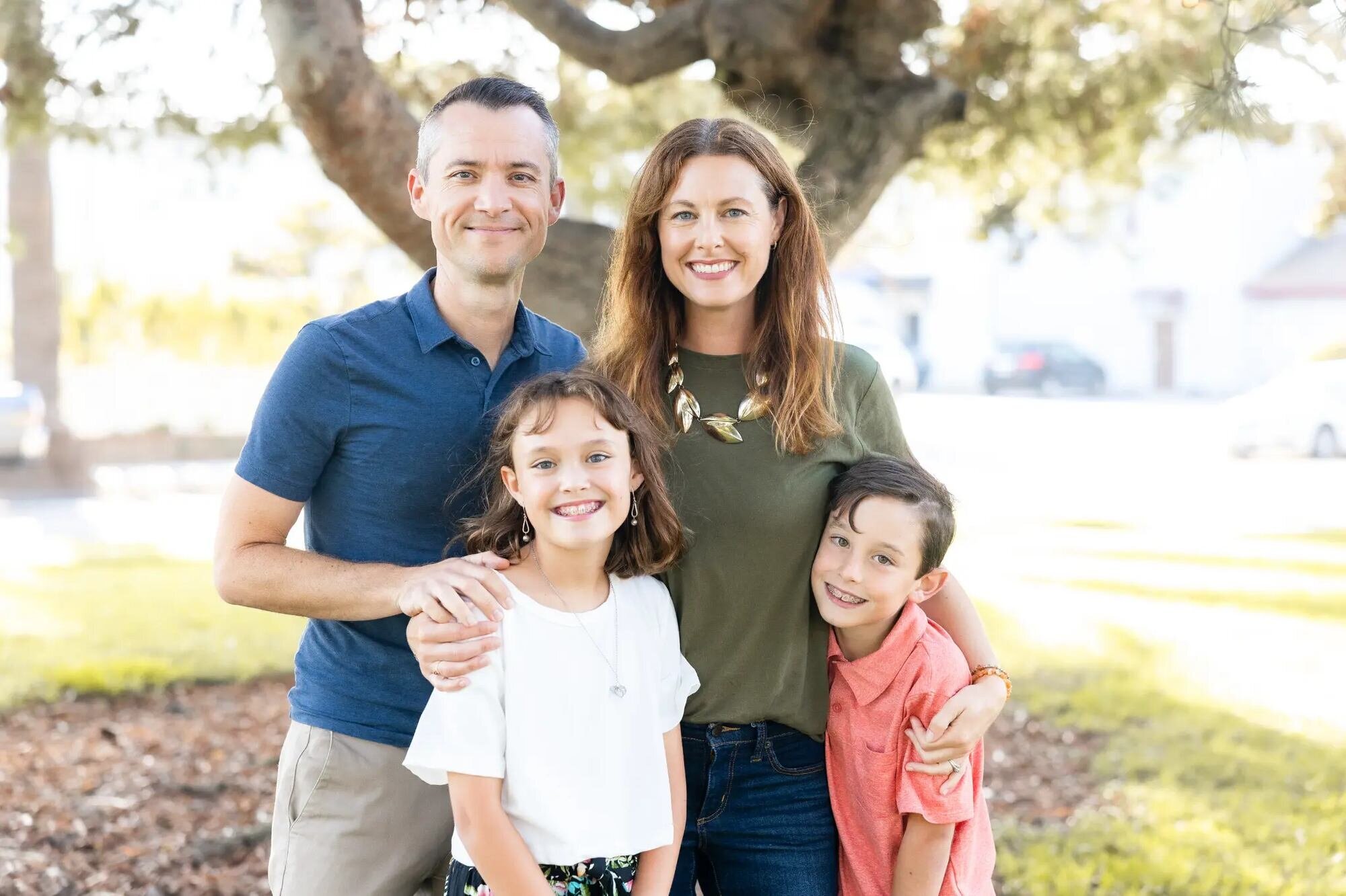 Los Angeles family photographer capturing a family of four with two school-age children smiling together under a large oak tree at Seal Beach in warm afternoon light