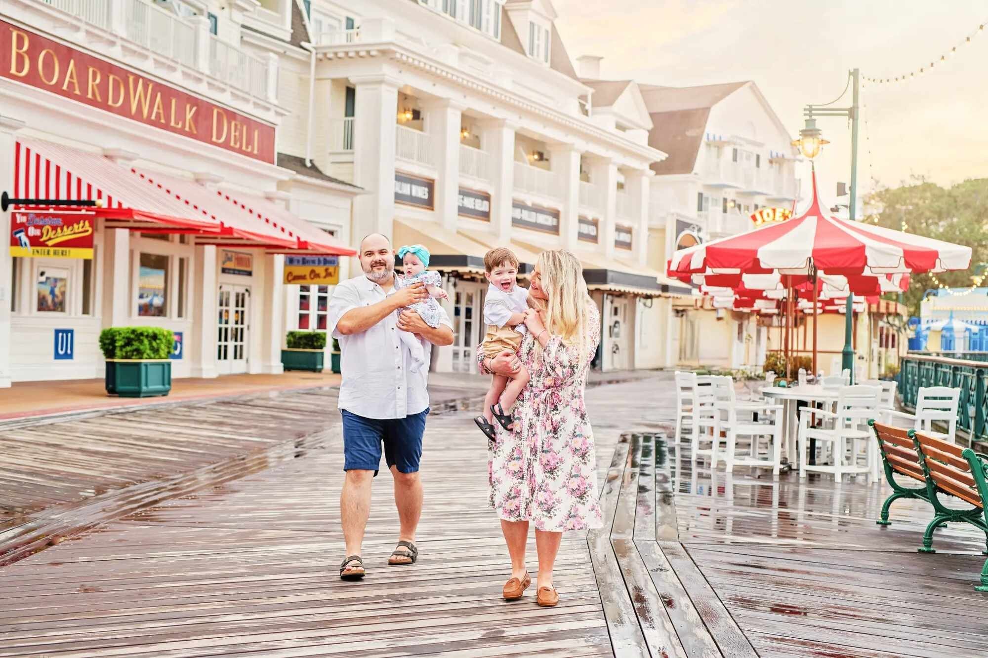 A family of four walking and laughing on the wooden promenade at Disney's BoardWalk Resort in Orlando, with colorful resort storefronts and warm golden light in the background