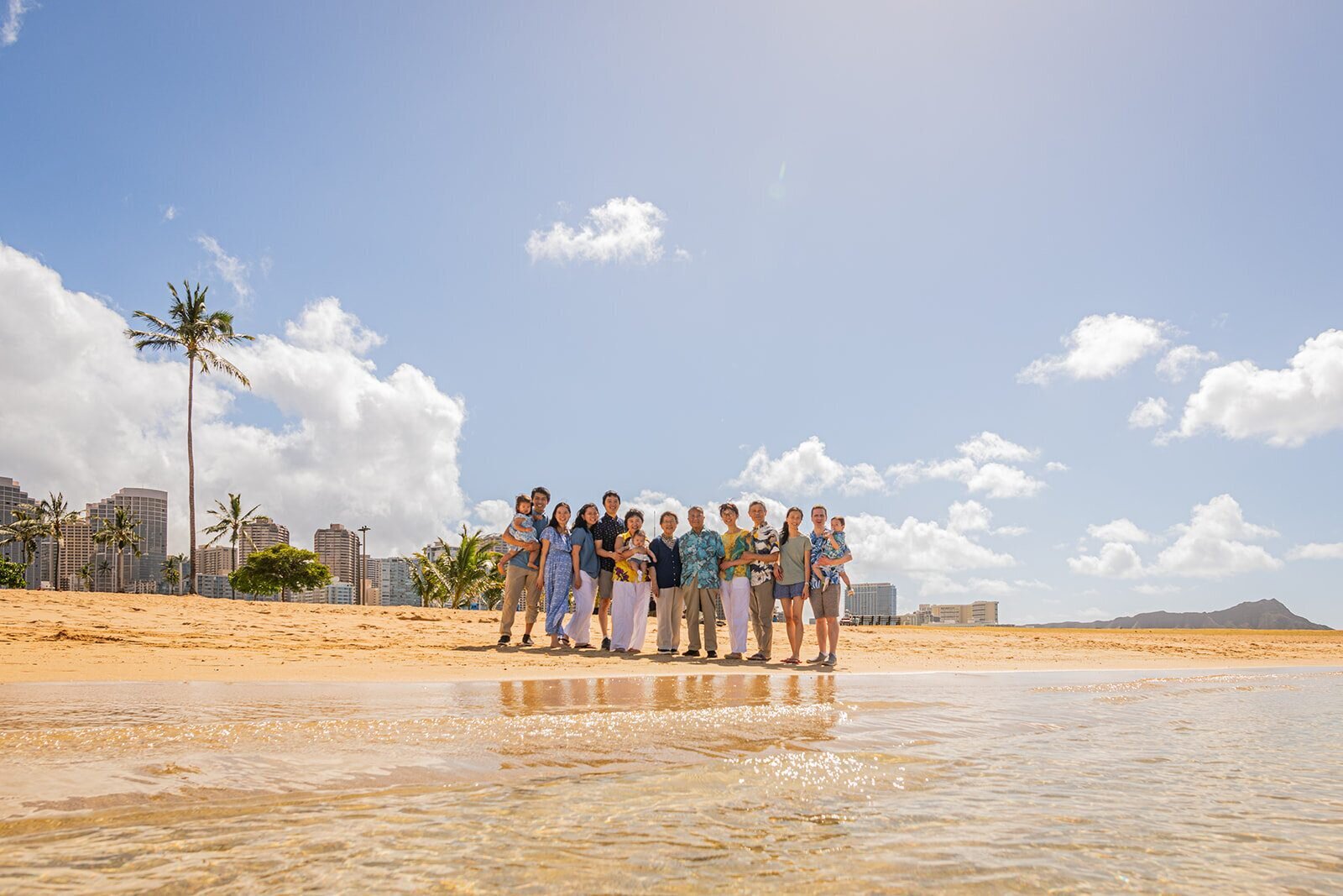 A large extended family of twelve posing together on the sand at Ala Moana Beach Park on Oahu with the Waikiki skyline, a tall palm tree, and Diamond Head visible in the background
