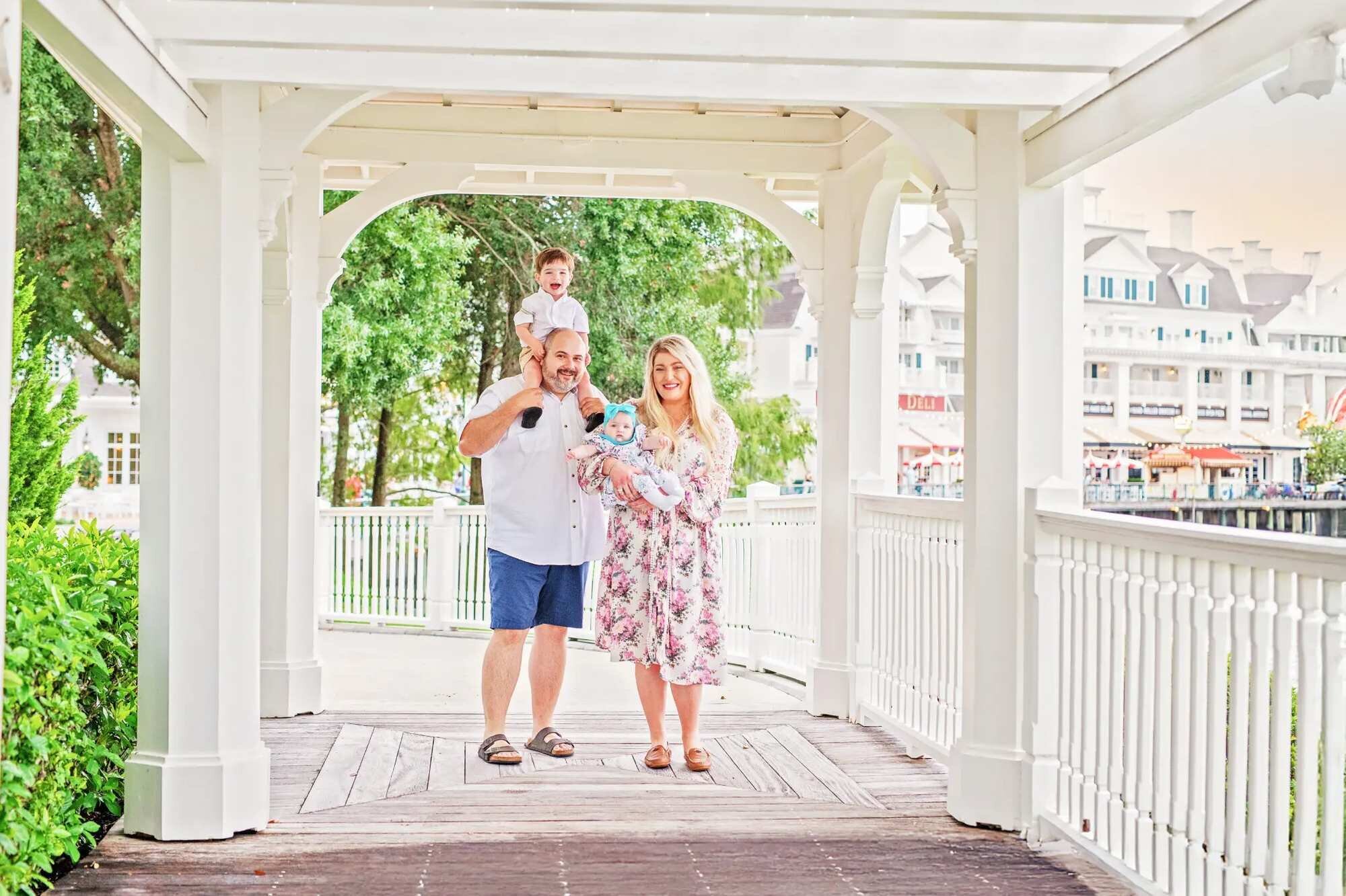 A family of four posing and laughing inside a white gazebo walkway at Disney's BoardWalk Resort in Orlando, with a dad carrying a toddler on his shoulders and a mom holding a baby, with the resort's elegant white architecture visible in the background