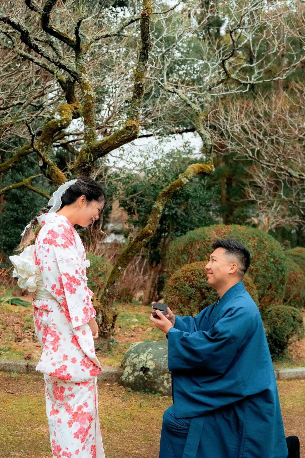 A man proposing on one knee in traditional blue kimono to his partner dressed in a floral furisode, surrounded by moss-covered trees in a Japanese garden in Fukuoka. A beautifully intimate proposal moment captured by Dreamgraphy.