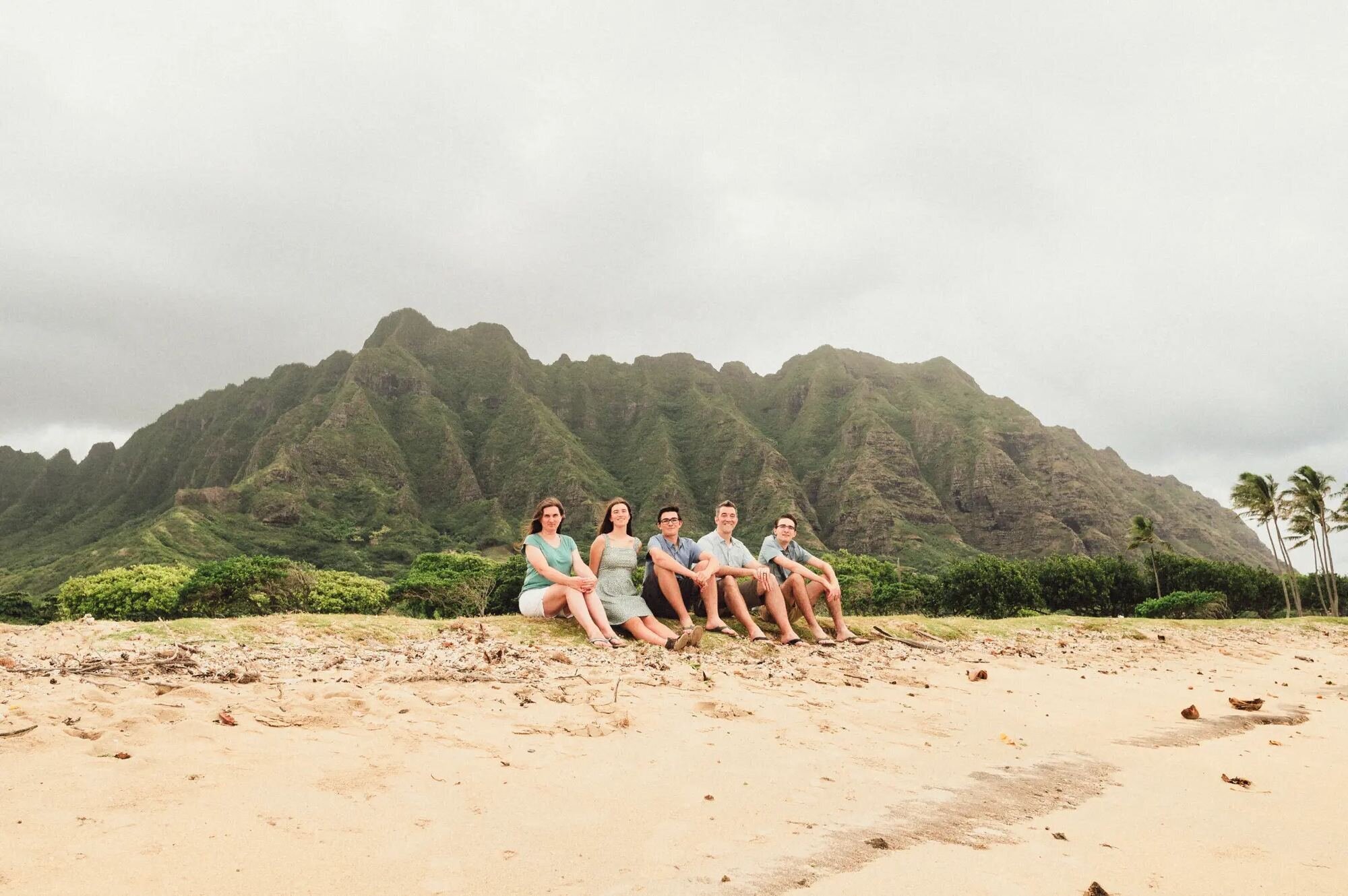 Oahu Photographer — Family Session at Kualoa Beach with Dramatic Hawaii mountains A family of five sitting together on the sand at Kualoa Beach on Oahu with the dramatic green Ko'olau mountain cliffs rising behind them