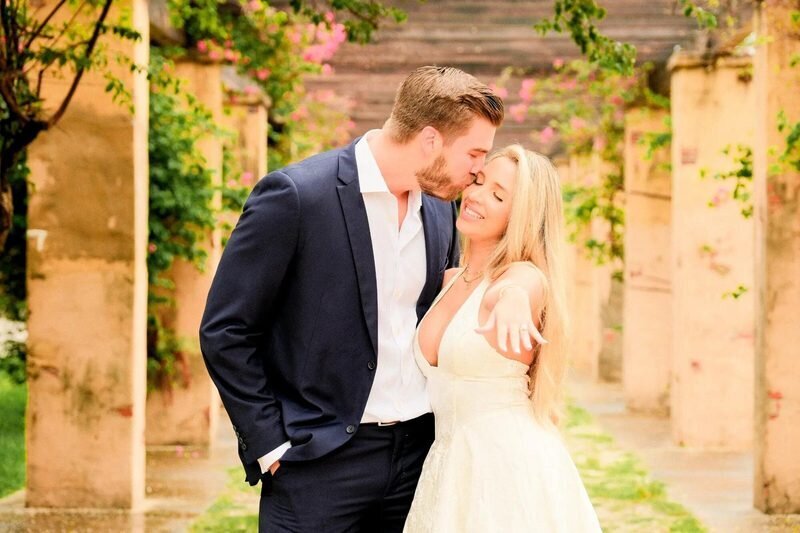 A man in a navy suit kissing the cheek of a smiling woman as she shows her ring, among stone pillars and pink bougainvillea in Miami