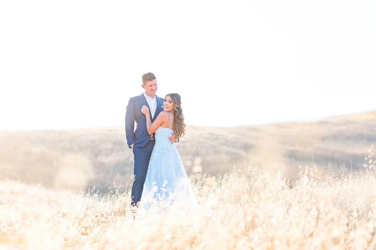 Los Angeles Photographer — Engagement Session at Malibu Creek State Park A man in a navy suit and a woman in a pale blue gown embracing in tall golden grass on a sunlit hillside in Los Angeles