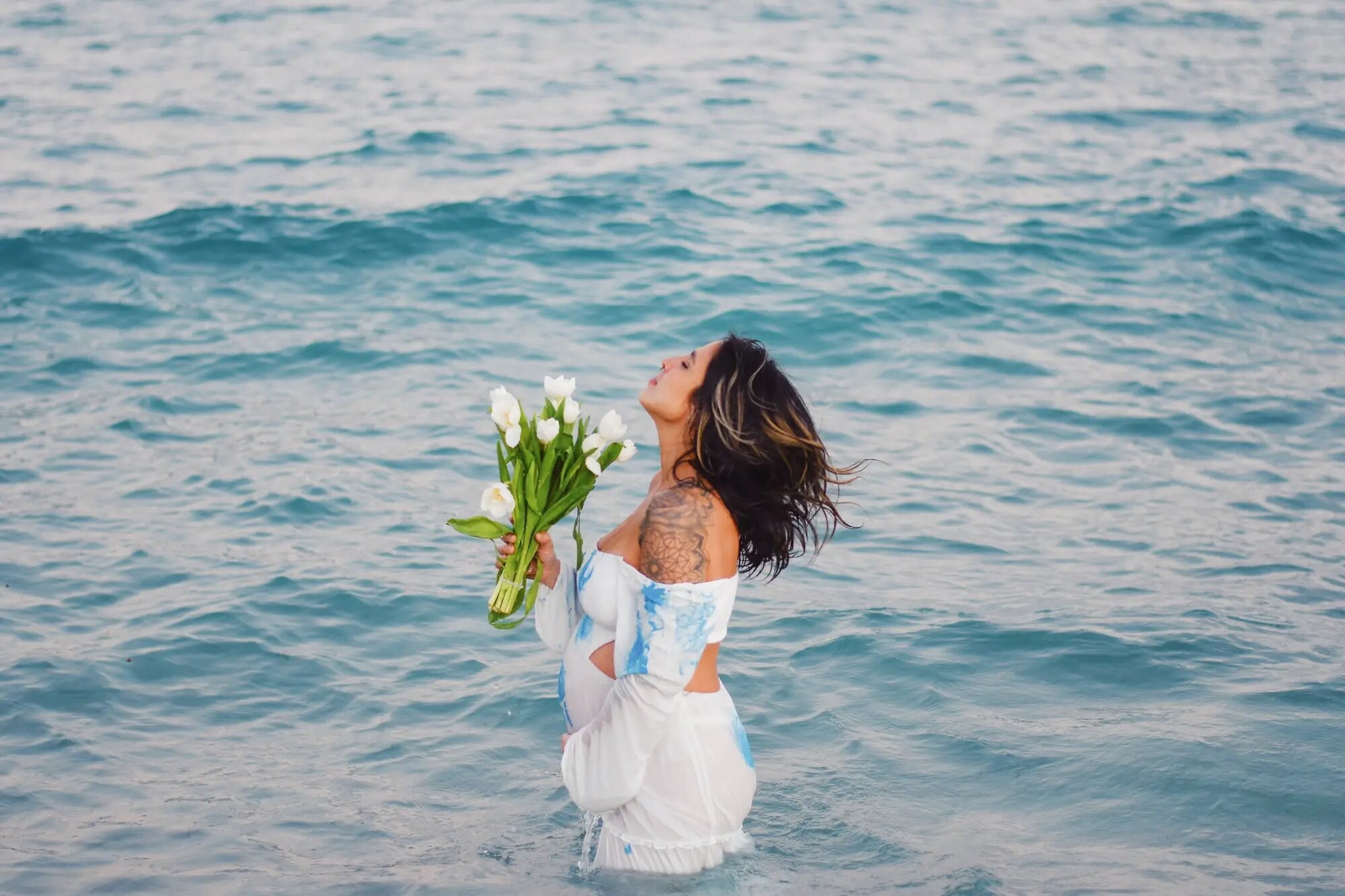 A pregnant woman standing in the turquoise ocean at South Beach, Miami, holding a bouquet of white tulips with her head tilted back — captured by a Dreamgraphy Miami maternity photographer