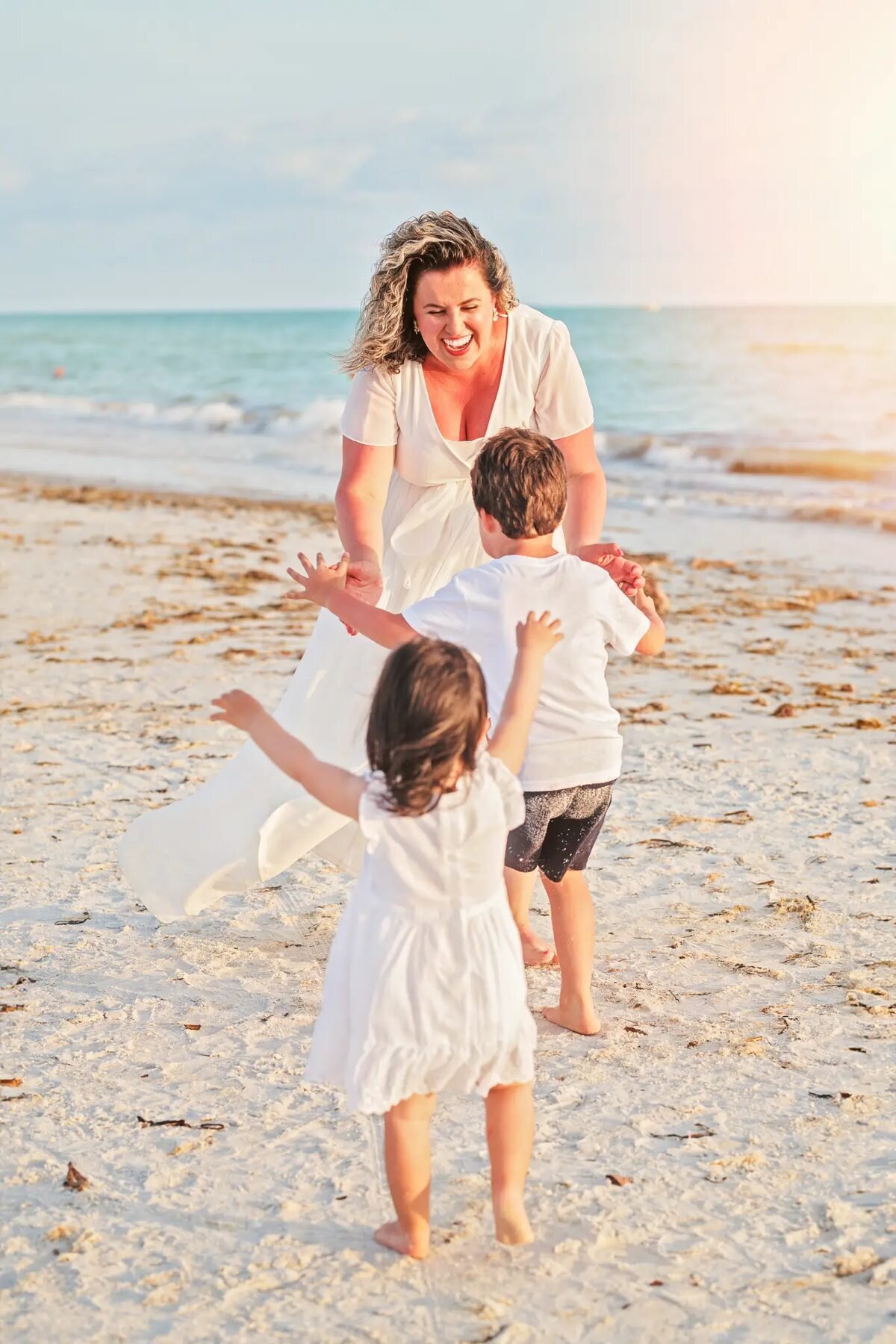 A mom laughing as her two young children run toward her on a Florida beach at golden hour, captured by a professional Orlando family photographer