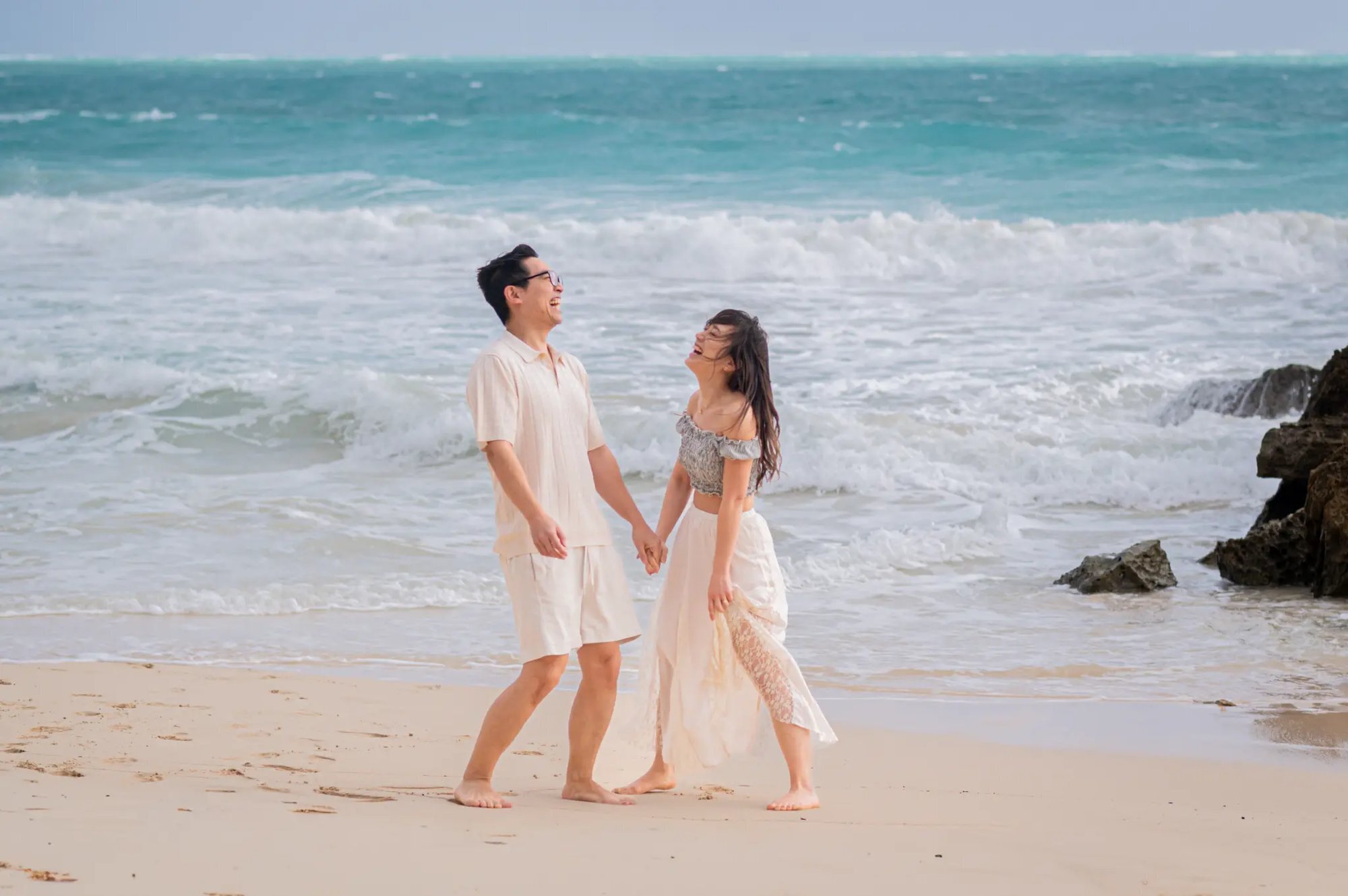 A couple holding hands and laughing together while walking along a sandy Kauai beach with turquoise ocean waves crashing behind them, captured during a professional couples photo session