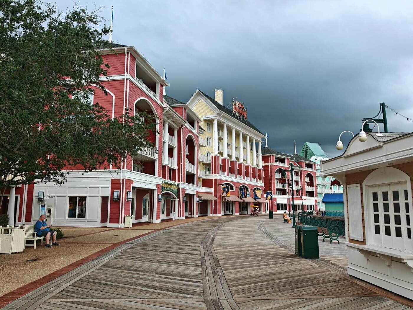 A curving wooden boardwalk past red and yellow resort buildings with white columns and green lamp posts under a stormy sky in Orlando