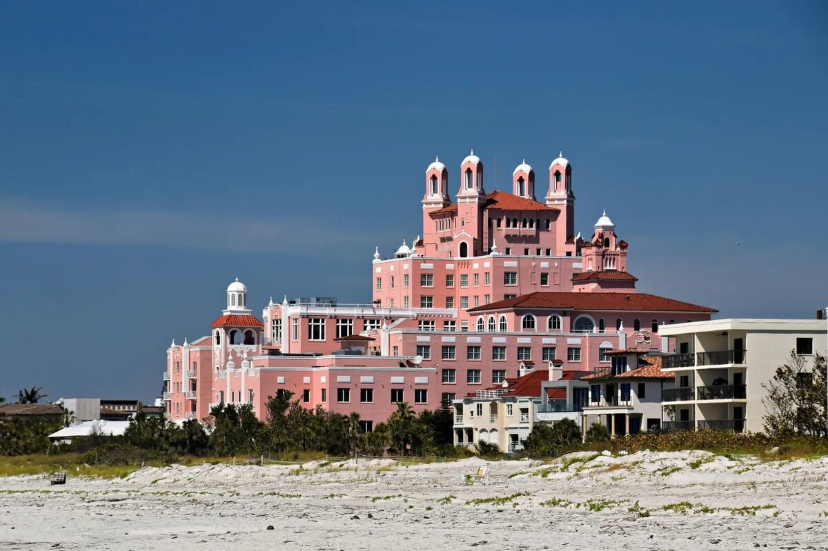 A large pink hotel with white-domed towers and red tile roofs rising behind white sand dunes under a deep blue sky in Tampa