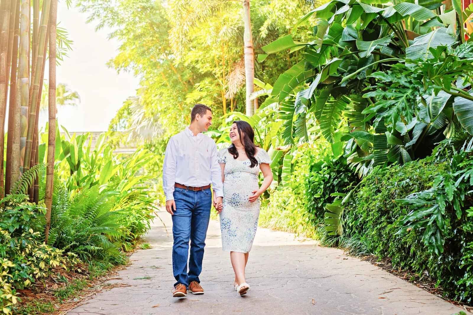 A smiling pregnant woman holding her belly and walking hand in hand with her partner along a lush tropical garden pathway in Orlando, Florida, surrounded by bamboo, banana leaves, and vibrant green foliage in warm golden light