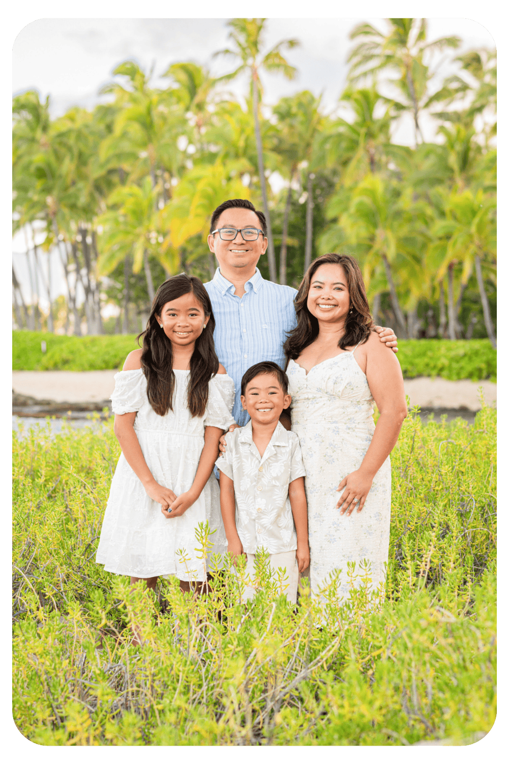 A family of four wearing coordinated white and light blue outfits smiling together among tropical greenery with palm trees on a Hawaii beach