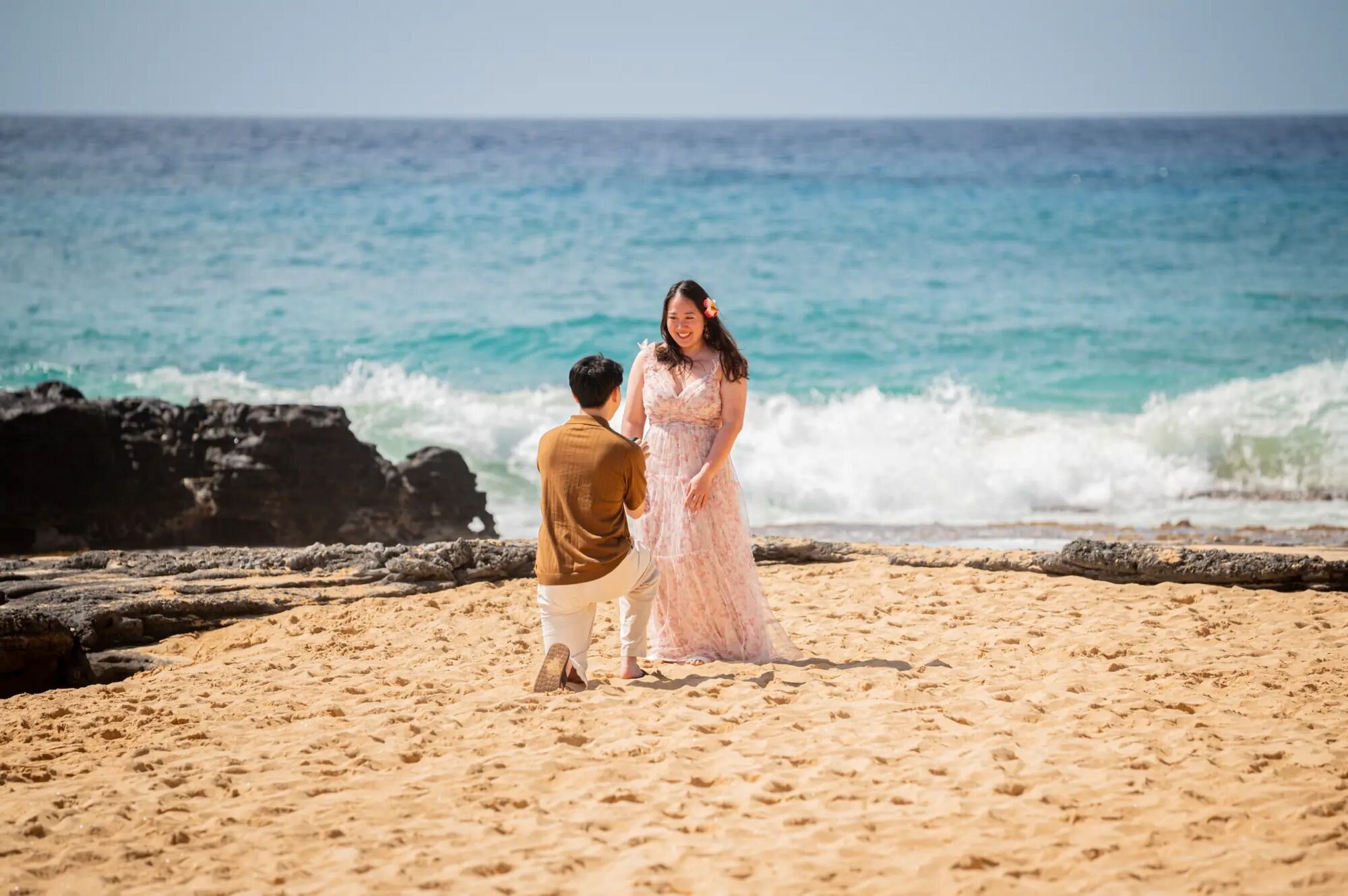 A man proposing on one knee on a sandy Kauai beach as his partner reacts with joy, with turquoise ocean waves and volcanic rocks in the background, captured by a professional Kauai proposal photographer